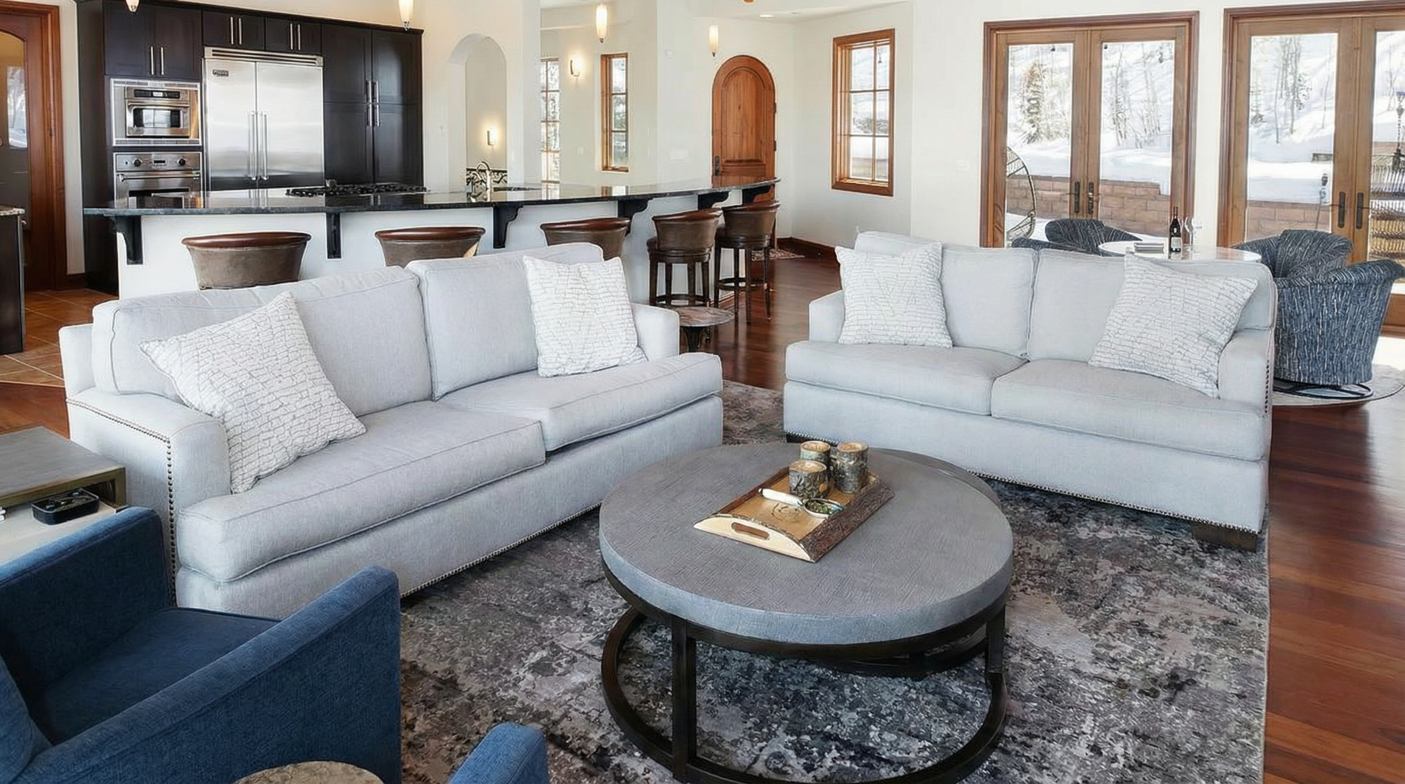 The living room features two light grey sofas, a round coffee table, and a blue armchair, all arranged on a patterned area rug. The open-concept design connects the living area to the kitchen and bar, creating a spacious and inviting atmosphere. The room's warm hardwood floors and natural light enhance the cozy ambiance.