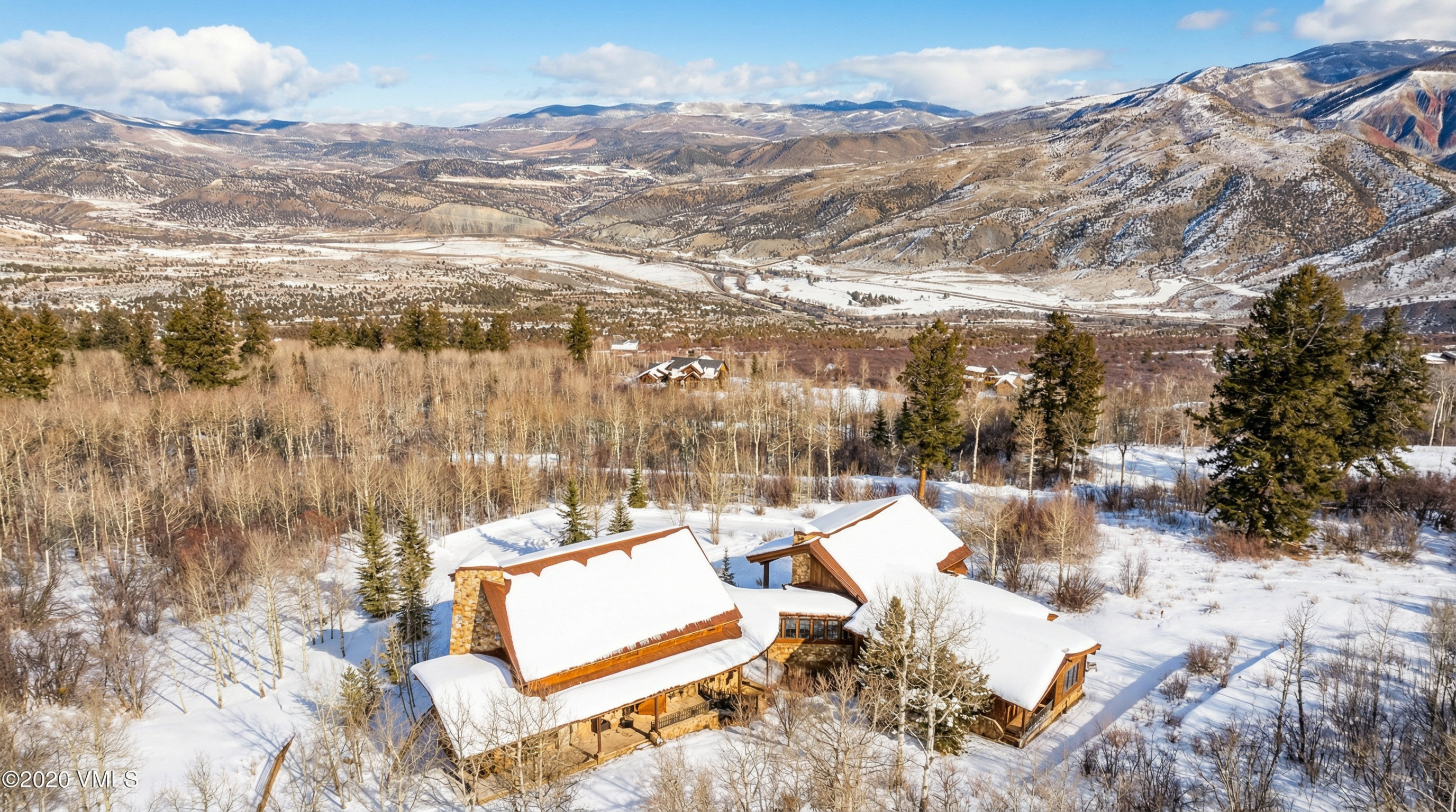 This aerial view captures a stunning mountain estate in winter. The snow-covered roofs of the rustic stone and wood home blend with the surrounding landscape of bare trees and snowy ground. The picturesque backdrop includes distant mountains and a clear blue sky, showcasing the property's secluded and scenic location.