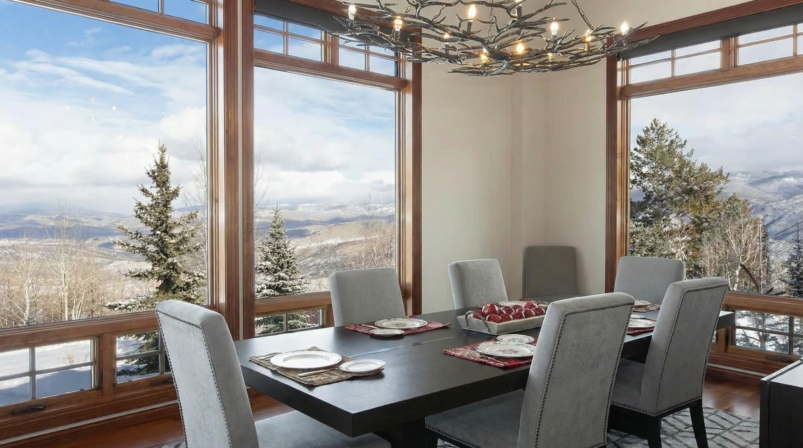 This interior shot showcases a dining room featuring a dark wooden table set for a meal, surrounded by six upholstered chairs. Large windows provide stunning views of a snow-covered mountain landscape, enhancing the dining experience. The room is illuminated by a unique, branch-like chandelier, adding an artistic touch to the sophisticated and serene atmosphere.
