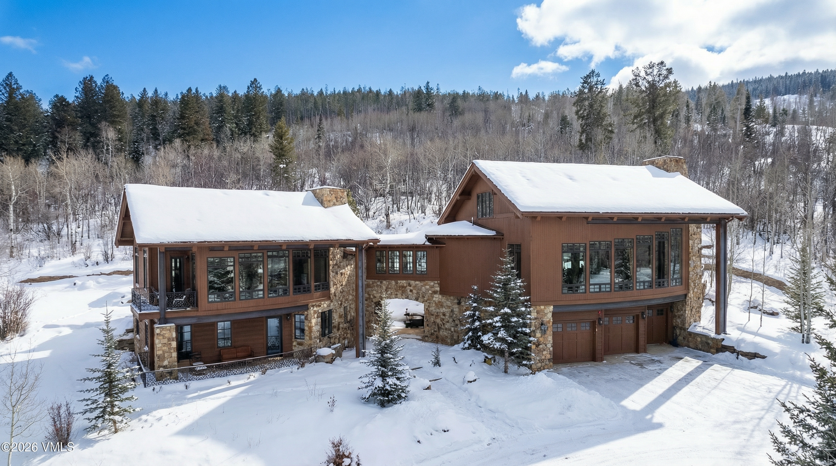 This aerial view captures a luxurious mountain home in a snowy landscape. The property features two main structures connected by a stone archway, showcasing impressive architectural design. Snow-covered roofs and surrounding evergreen trees contribute to a picturesque winter scene.
