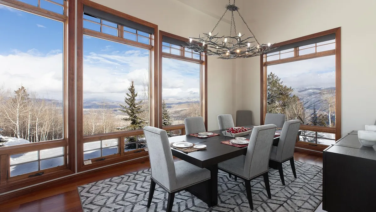 This is an interior shot of a dining room featuring large windows with an expansive mountain view. The dark wood dining table is set for a meal, and the six upholstered chairs provide comfortable seating. An elaborate chandelier hangs above the table, adding an elegant touch to the space.