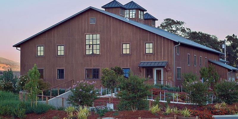 This image showcases a charming, contemporary barn-style building with vertical board-and-batten siding and a prominent gabled roof featuring cupolas. The structure includes multiple multi-pane windows and a welcoming entryway marked by a metal-roofed awning. Lush landscaping, including varied shrubs and flowering plants, enhances the curb appeal, creating a serene and inviting atmosphere.