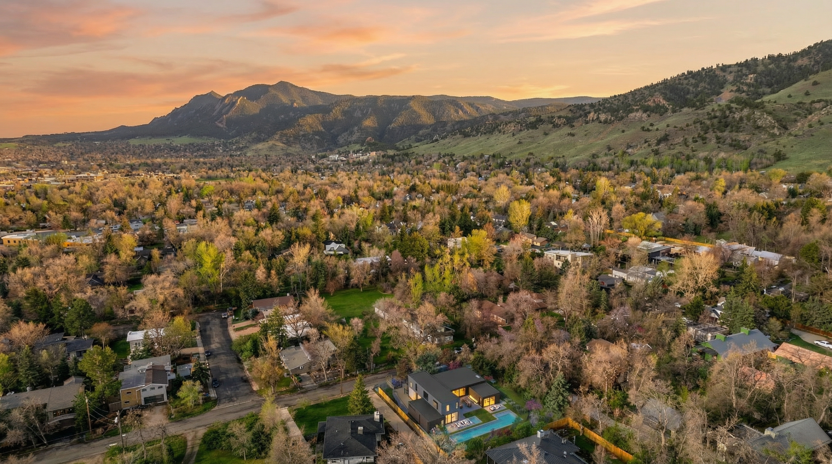 This aerial view captures a modern, multi-level residence set within a lush, tree-filled neighborhood against a stunning backdrop of the Rocky Mountains. The property features a contemporary architectural design, a swimming pool, and a well-integrated outdoor living space. The soft, warm lighting of the sunset enhances the peaceful and upscale atmosphere of this highly desirable location.