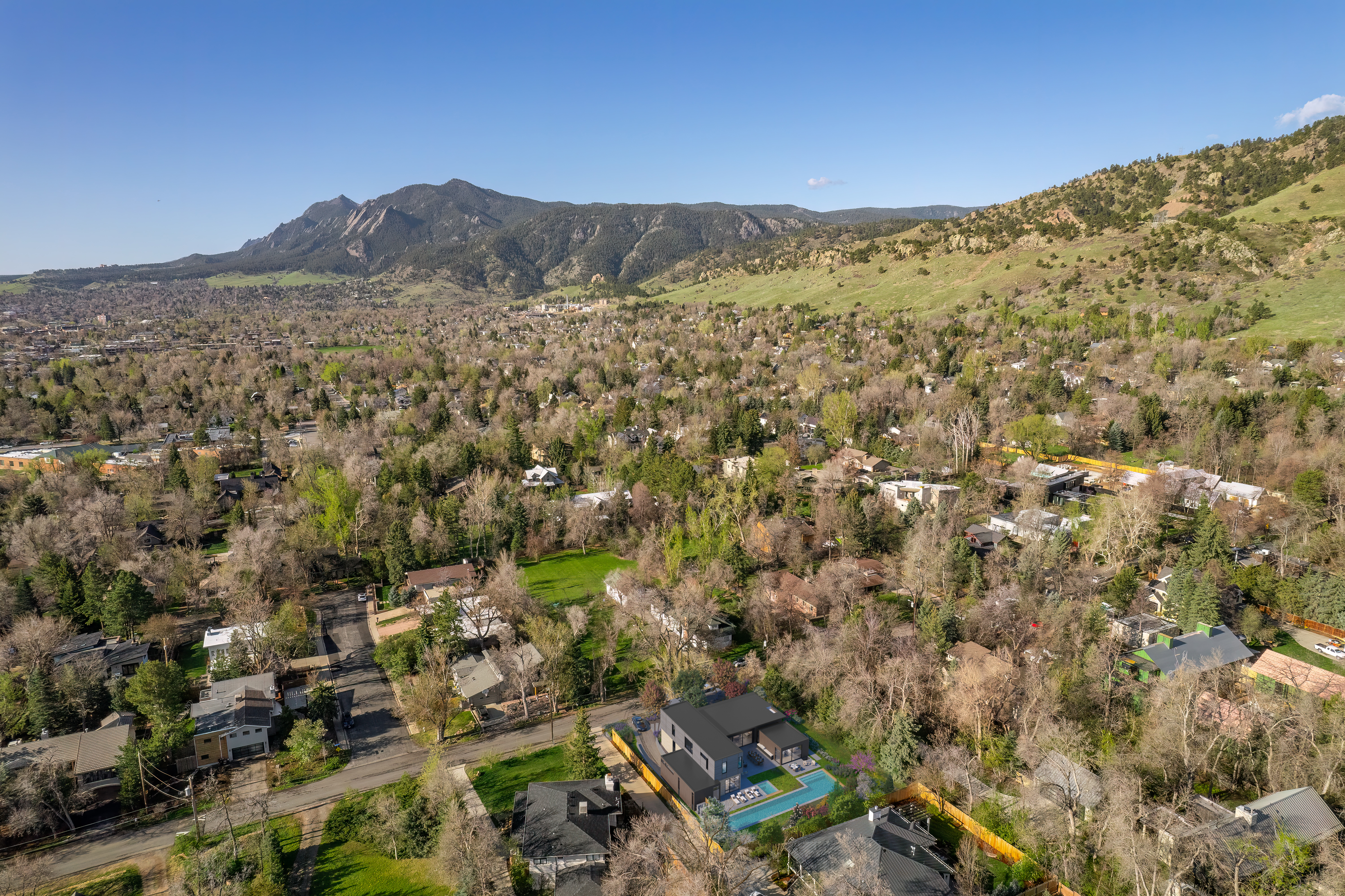 This is an aerial perspective showcasing a neighborhood nestled against a mountainous backdrop. The scene highlights a newly integrated modern residence featuring a dark, sleek design with an expansive outdoor pool area. The development blends contemporary architectural elements into an established, tree-lined residential community.