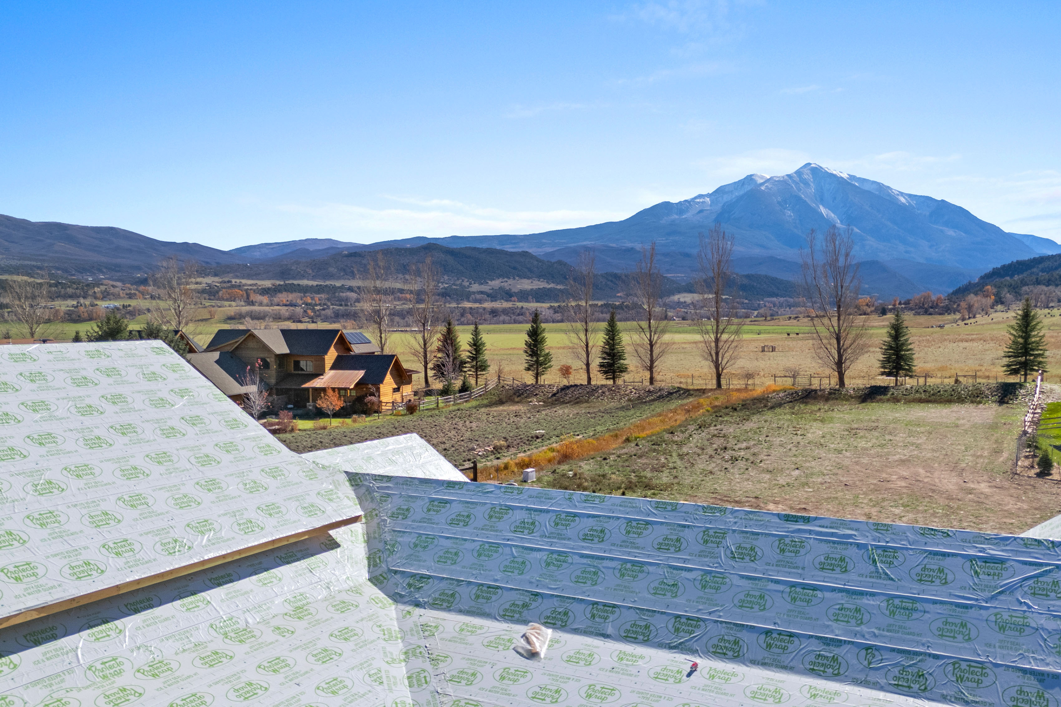 This elevated perspective captures a house under construction, with a focus on the newly applied roofing underlayment in the foreground. The property overlooks a vast, scenic rural landscape featuring rolling fields, mature trees, and majestic snow-capped mountains in the distance. The view highlights the home's prime location and picturesque surroundings, offering a blend of ongoing development and natural beauty.