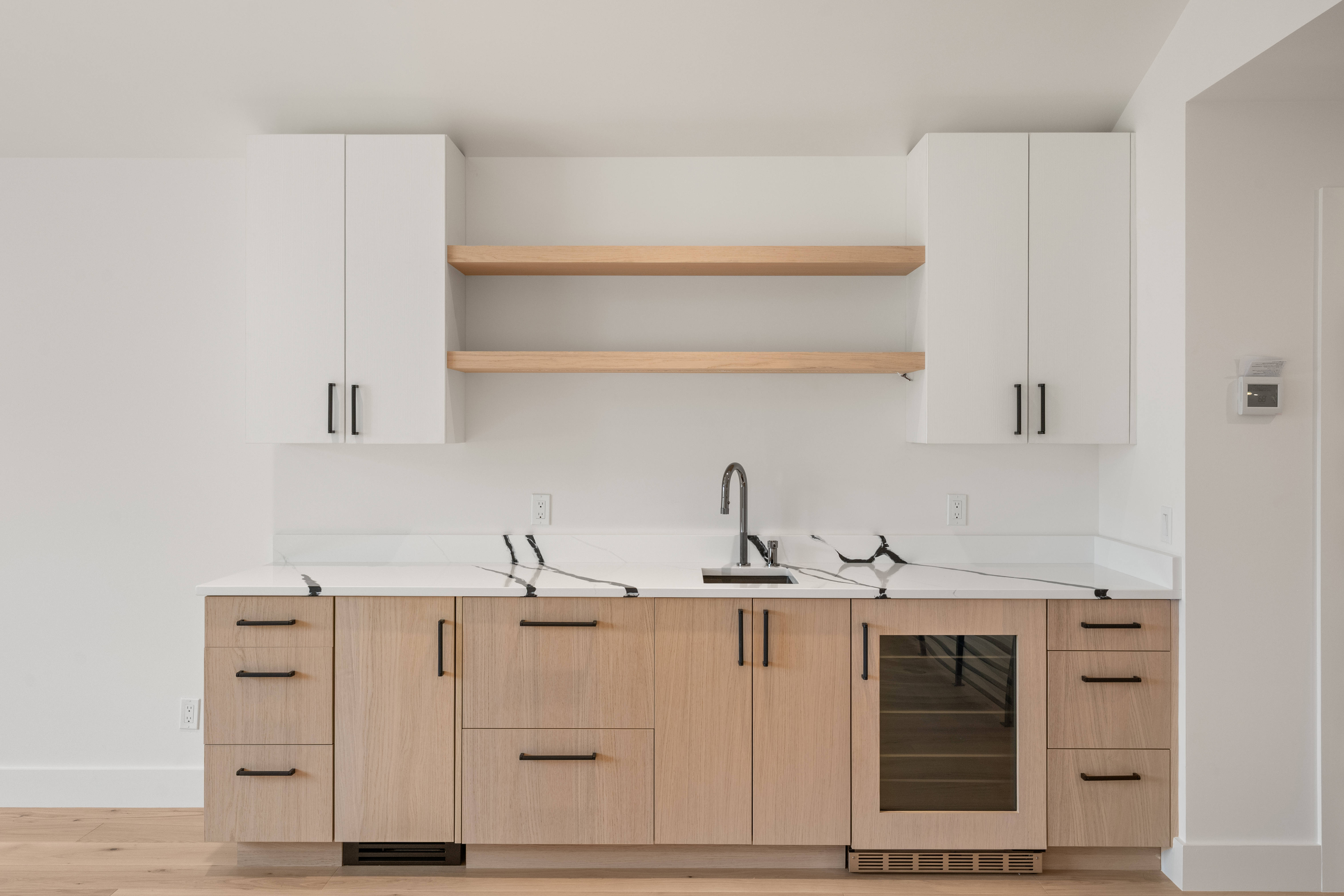 This modern kitchenette features light wood cabinetry accented by white upper cabinets and elegant marble-patterned countertops. It is equipped with a functional sink, integrated wine cooler, and floating wooden shelves, creating a sleek and minimalist aesthetic. The space is well-lit and designed for both utility and contemporary style.