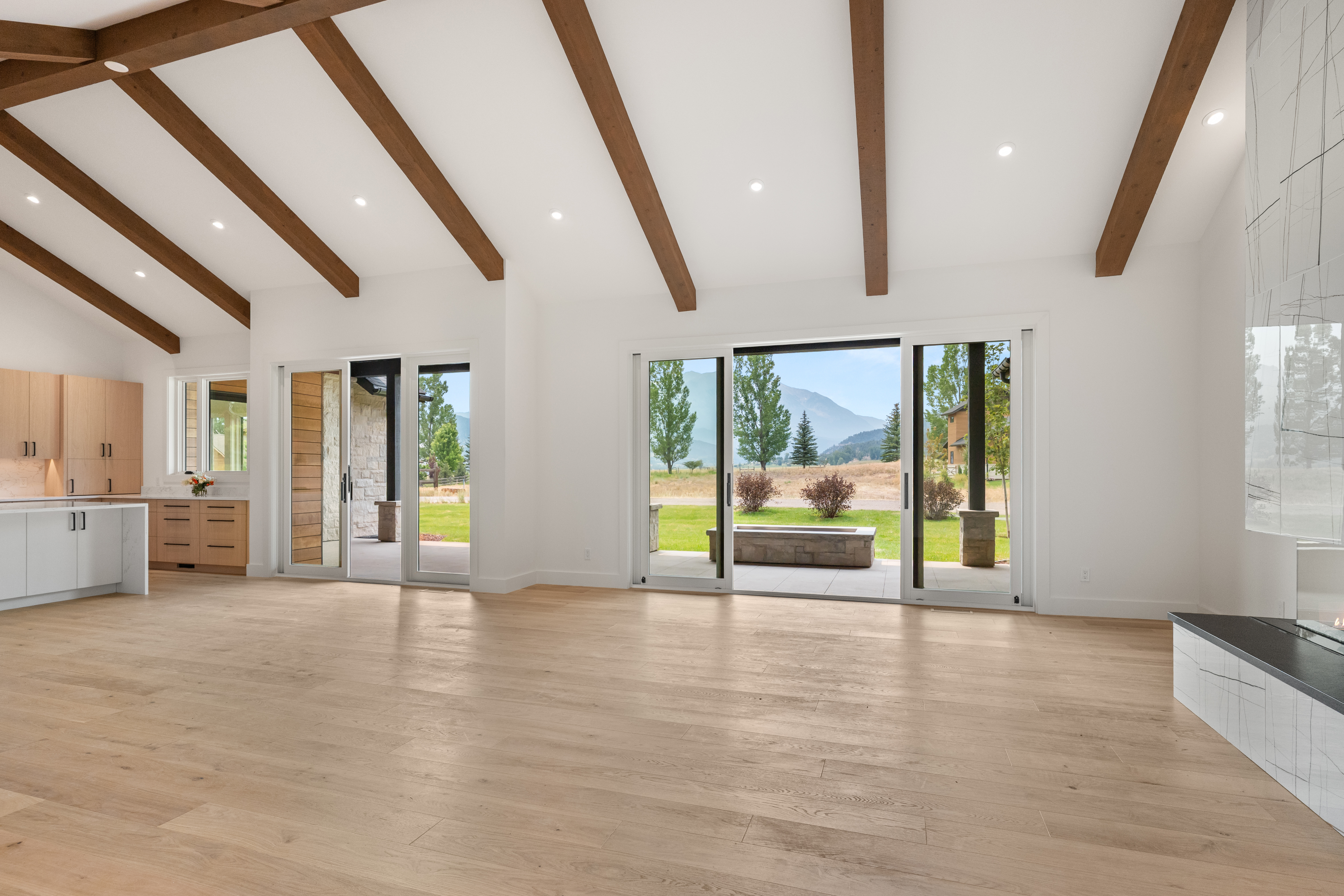 This bright and spacious living area features high vaulted ceilings with exposed wood beams and light hardwood flooring, creating an airy, modern aesthetic. Large sliding glass doors provide a seamless connection to the outdoor patio and offer stunning views of the surrounding mountains. The room is open-plan, flowing directly into a sleek kitchen, making it perfect for both relaxation and entertaining.