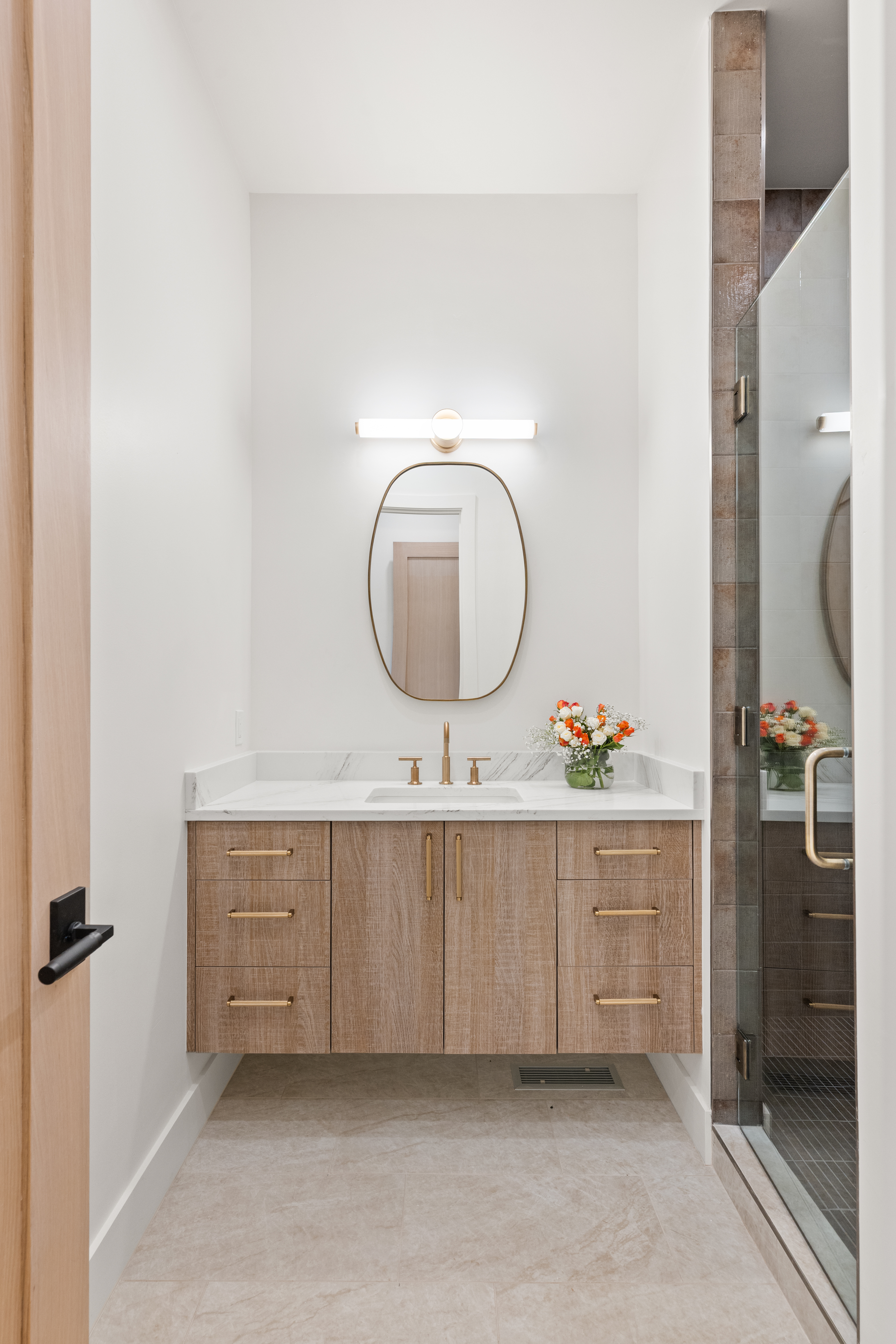 This modern guest bathroom features a sleek floating wood vanity with gold hardware, topped with a clean marble countertop and an undermount sink. A contemporary oval mirror and a minimalist light fixture enhance the bright, airy aesthetic, while the adjacent shower area is enclosed by clear glass with tasteful tile work. The overall design is refined and sophisticated, offering a high-end feel perfect for a luxury property.