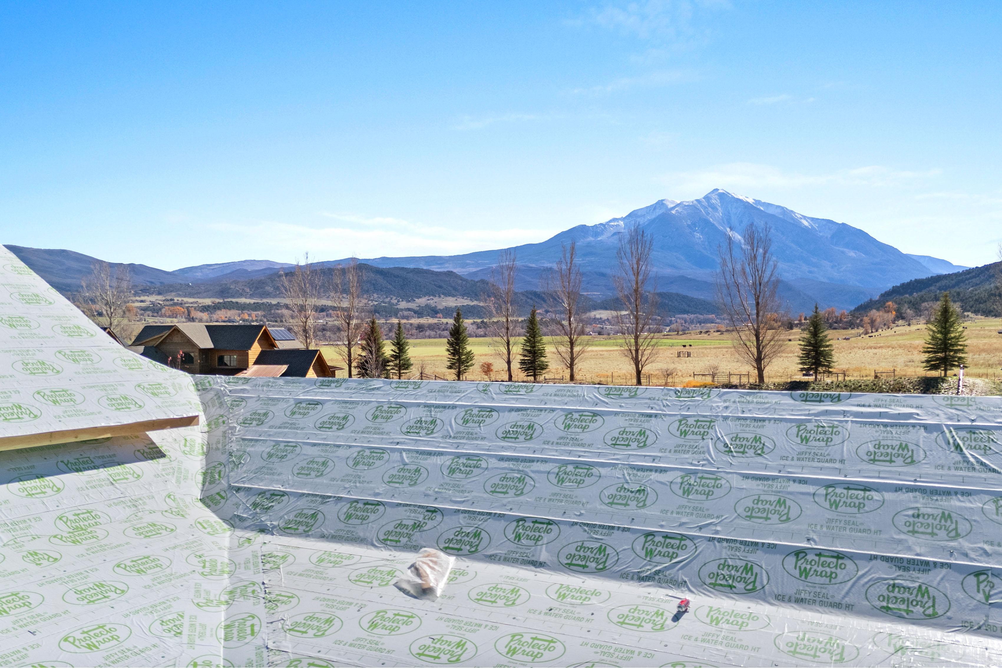 This image provides an elevated vantage point looking out over a rural landscape featuring a majestic snow-capped mountain in the distance. The foreground consists of a roof under construction, covered in protective waterproof membrane rolls, suggesting a new build or major renovation. The setting offers a picturesque, serene view of open fields, mature trees, and a neighboring mountain property.