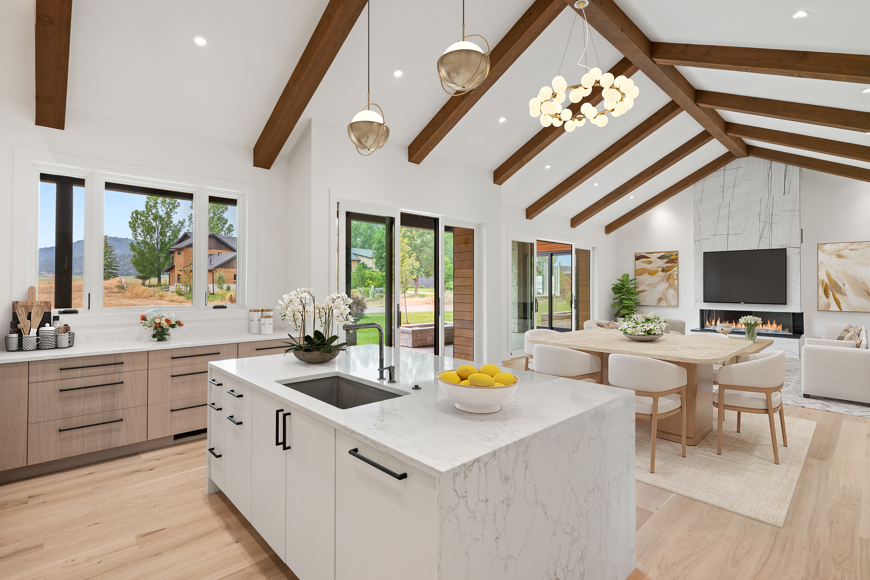 This bright and airy open-concept kitchen features a large white marble-topped island and light wood cabinetry, perfectly blending modern luxury with natural elements. High vaulted ceilings with exposed wood beams create a spacious feel, while seamless glass doors invite views of the scenic outdoor landscape. The space is complemented by a sophisticated chandelier, contemporary pendant lighting, and an adjacent dining area, making it an ideal setting for entertaining.