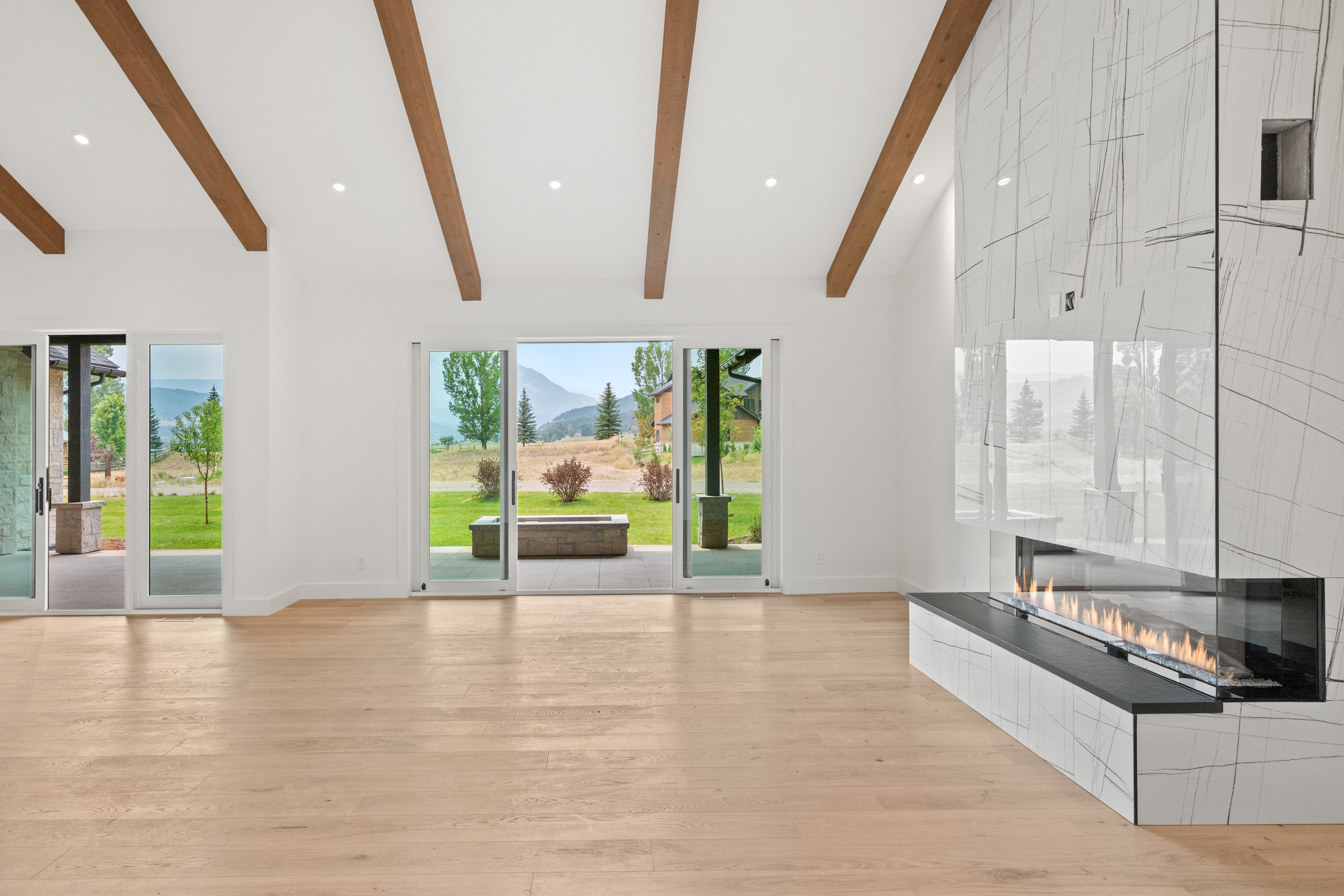 This spacious living area features a high vaulted ceiling with exposed wooden beams, creating an airy and open atmosphere. A stunning, modern floor-to-ceiling fireplace with white marble-style cladding serves as the focal point, while expansive sliding glass doors provide seamless indoor-outdoor transition and panoramic views of the mountain scenery. The minimalist design is complemented by warm hardwood flooring, resulting in a bright, sophisticated, and contemporary home aesthetic.