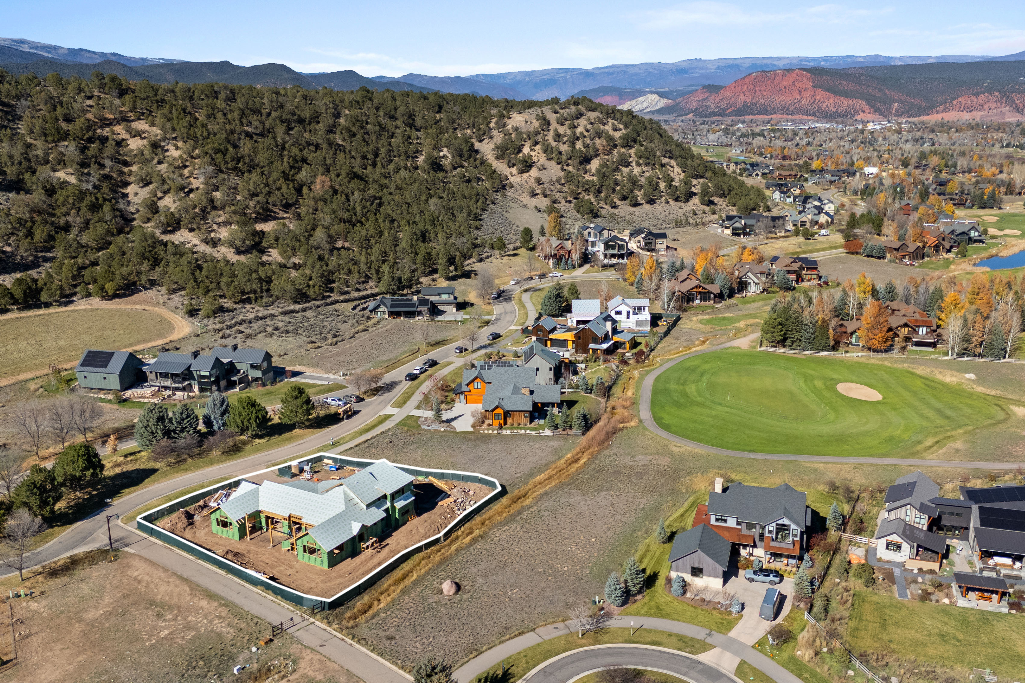 This high-altitude aerial view captures a scenic, upscale residential neighborhood nestled near rolling hills and a golf course. The community features a mix of modern architecture, large single-family homes, and a construction site for a new development, all set within a lush, rural landscape. The bright, clear daylight accentuates the expansive views and the well-integrated design of the community.
