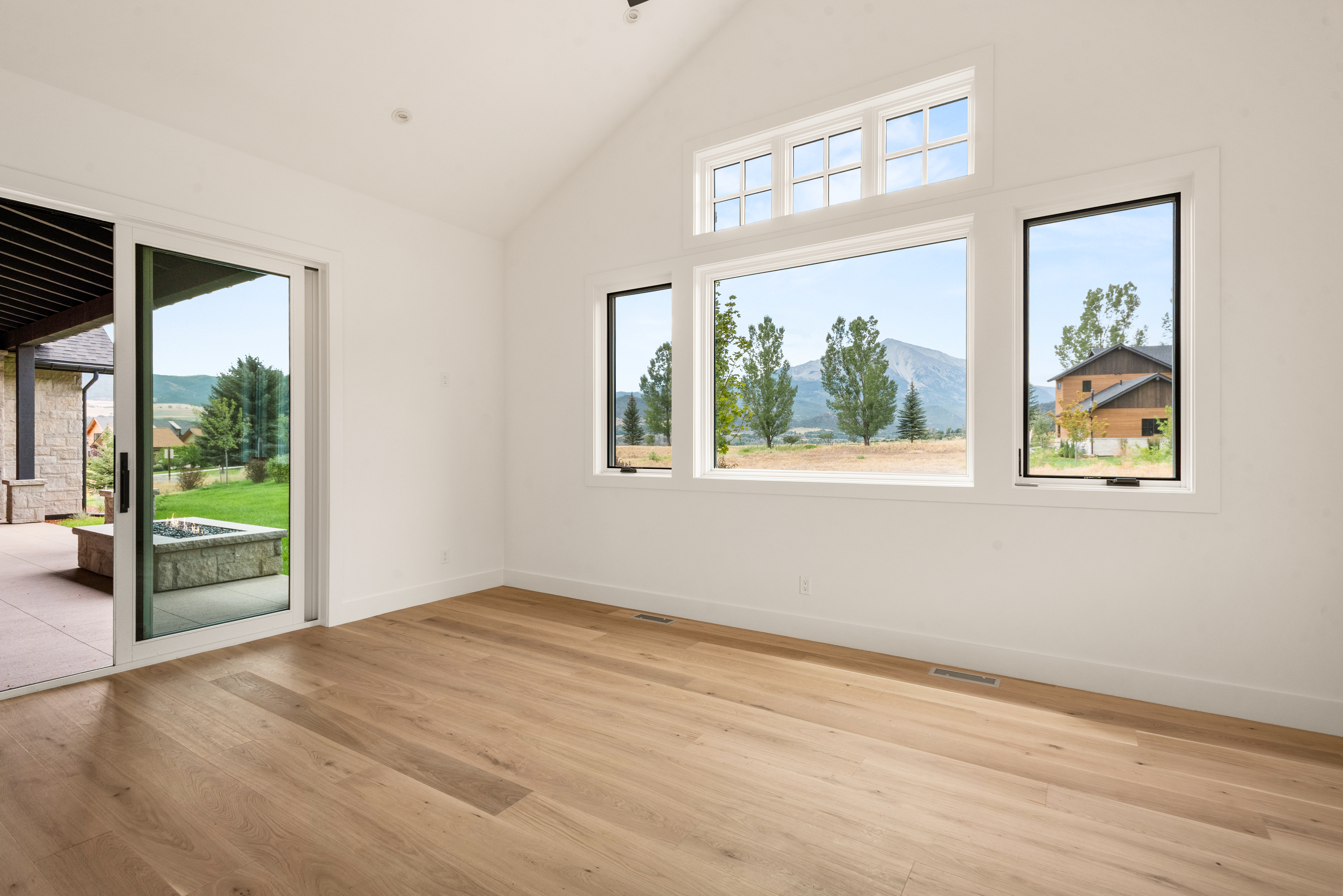 This spacious, modern living room features light-toned hardwood flooring, clean white walls, and vaulted ceilings that create an airy and open atmosphere. Large windows and a glass sliding door provide abundant natural light while framing picturesque mountain views and a patio area with a fire pit. The minimalist design and seamless indoor-outdoor transition make this an ideal space for comfortable living and entertaining.