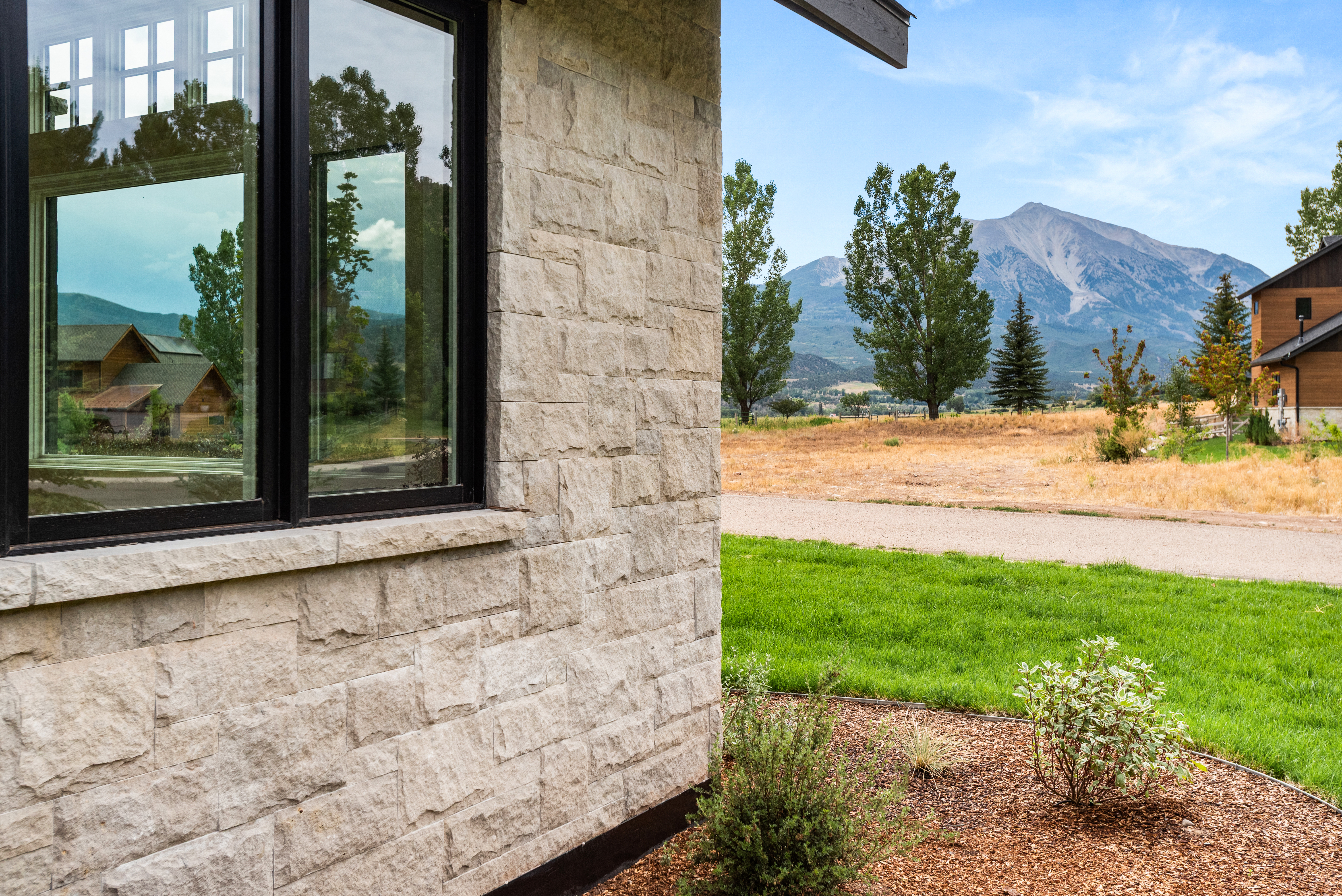 This exterior side view highlights a contemporary home featuring textured light-colored stone masonry and sleek black-framed windows. The scene captures a picturesque mountain backdrop over a well-maintained grassy yard, emphasizing the property's serene location and premium curb appeal. Reflective glass in the window provides a glimpse of the surrounding landscape, adding depth and architectural interest.