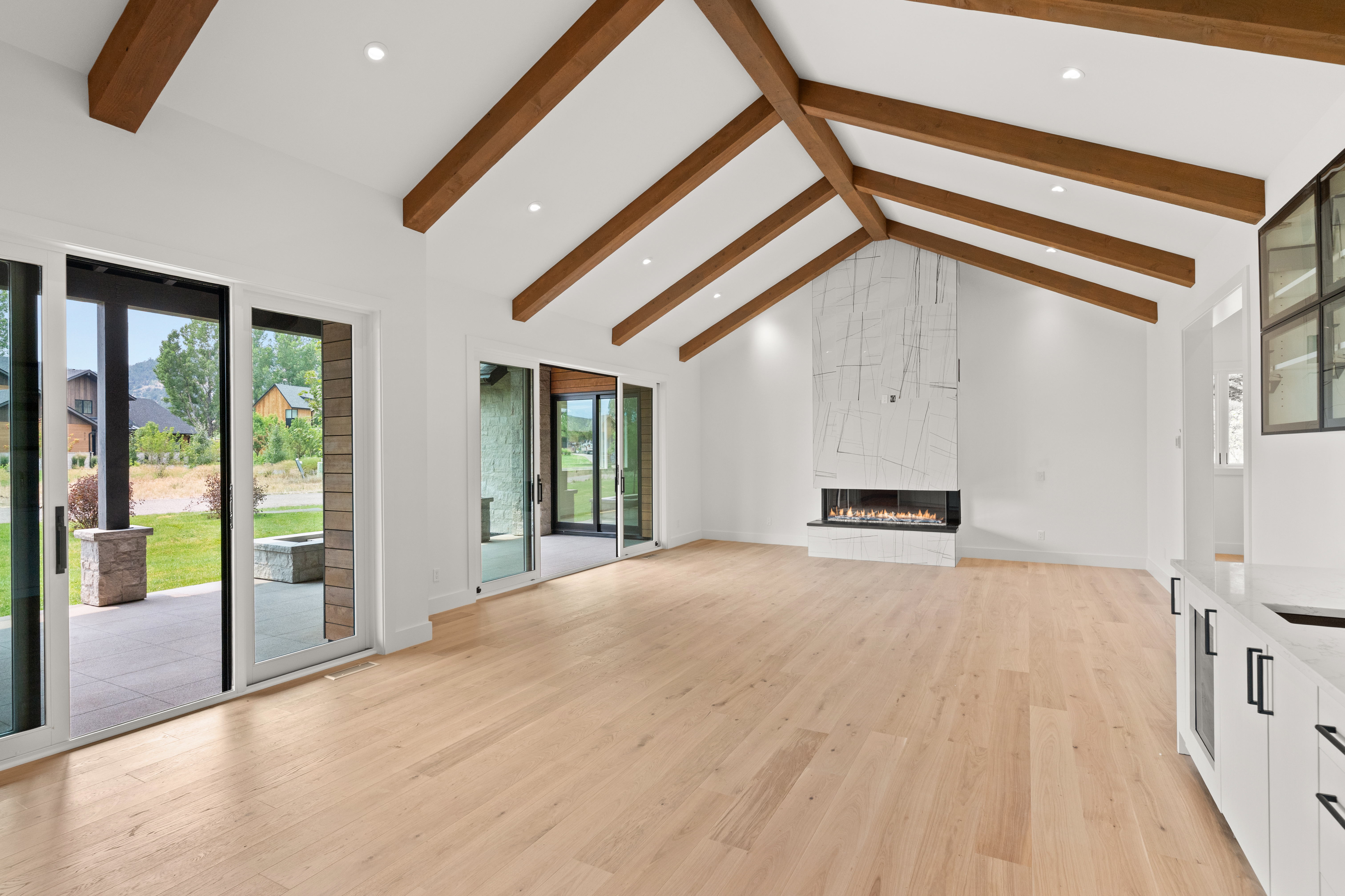 This spacious, open-concept living area features stunning vaulted ceilings with exposed wood beams and light hardwood flooring. The room is anchored by a contemporary fireplace with striking marble-patterned tile, and large sliding glass doors provide seamless indoor-outdoor flow to a patio. The bright, modern aesthetic is further enhanced by recessed lighting and high-end finishes, creating an inviting and airy atmosphere.