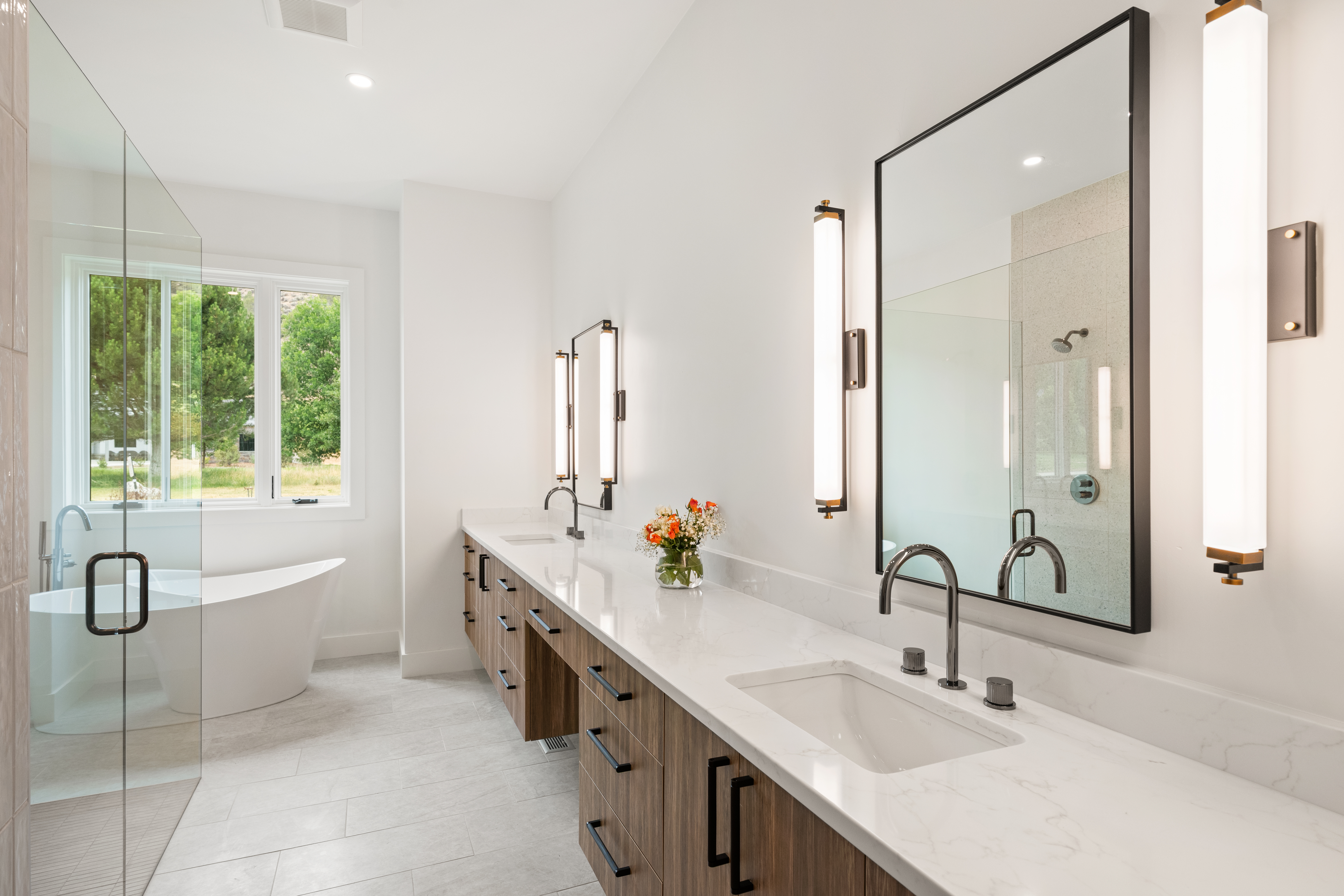 This luxurious primary bathroom features a sleek double vanity with wood cabinetry, marble-look countertops, and modern matte black fixtures. The space is bright and airy, highlighted by minimalist wall-mounted sconces, a spacious glass-enclosed shower, and a sophisticated freestanding soaking tub placed beneath a large window. Its clean lines and neutral color palette create a spa-like atmosphere suitable for a contemporary home.