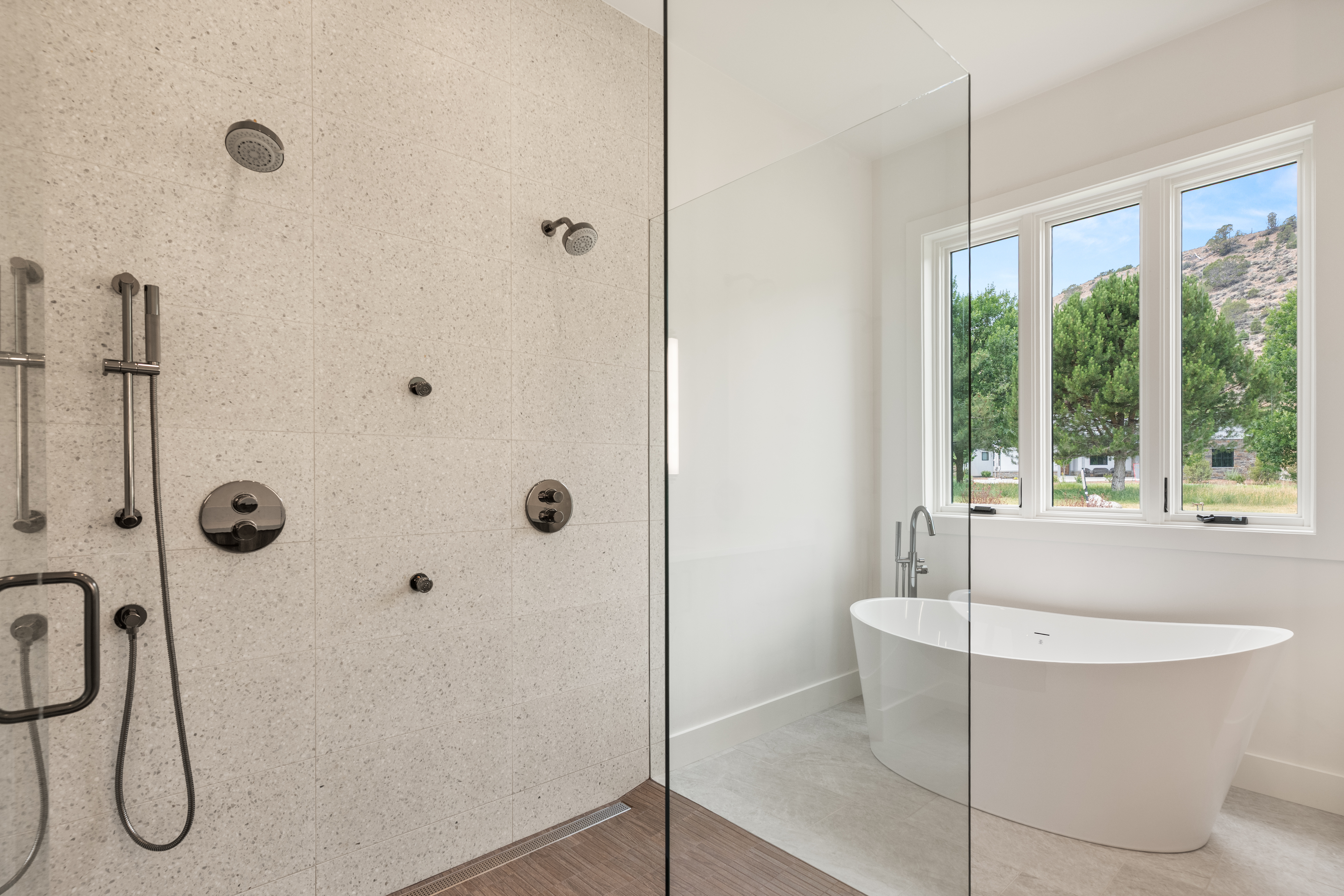 This modern bathroom features a spacious walk-in shower with high-end matte black fixtures, multiple shower heads, and speckled tile wall accents. Adjacent to the shower, a sleek, freestanding oval bathtub sits beneath a large window that fills the space with natural light and provides serene outdoor views. The combination of clean lines, neutral tones, and premium hardware creates a luxurious, spa-like atmosphere.
