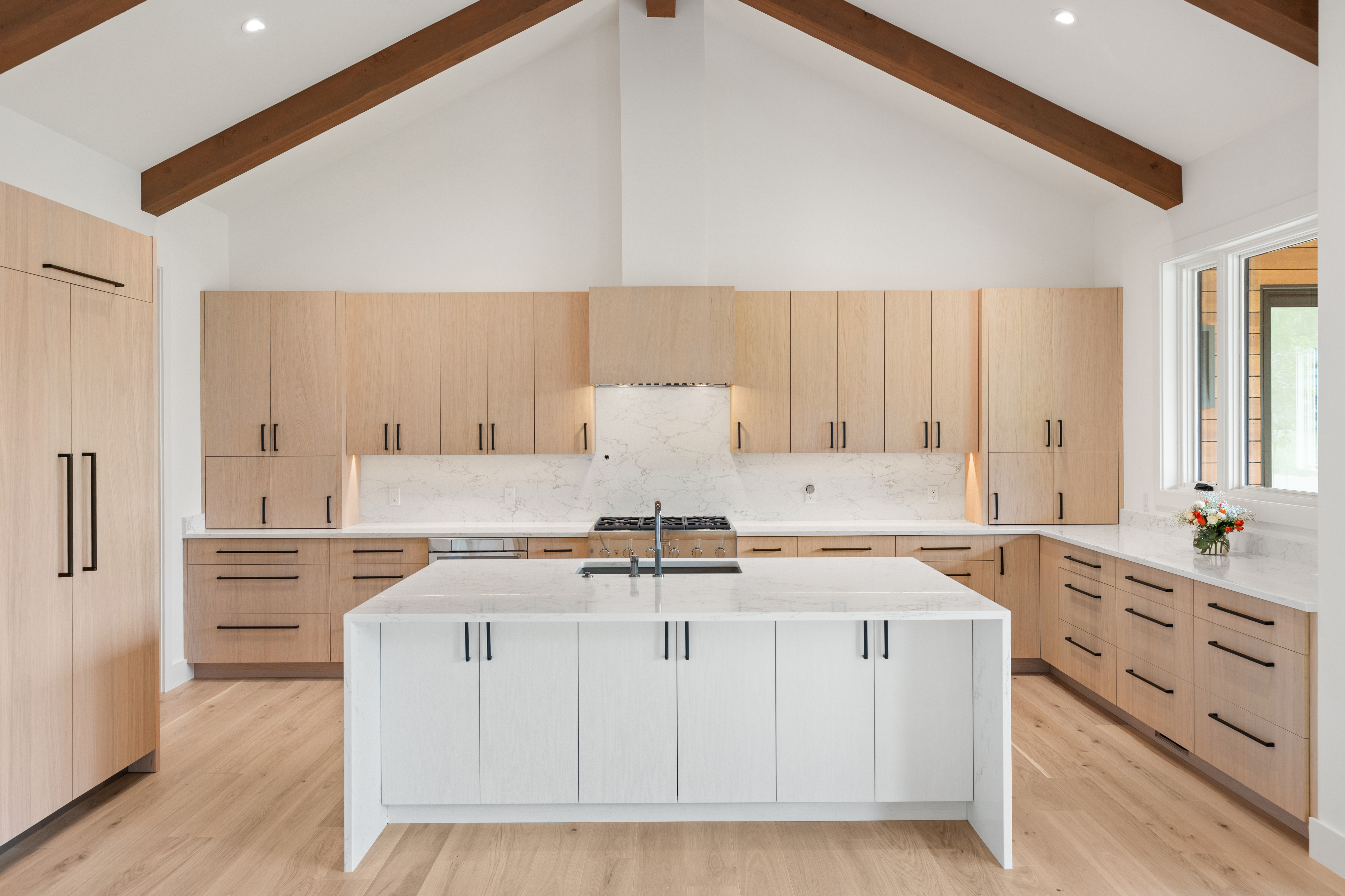 This modern kitchen features high-vaulted ceilings with exposed wooden beams, creating a spacious and airy atmosphere. The design combines light wood cabinetry with a crisp white kitchen island, complemented by sleek marble countertops and minimalist black hardware. Large windows and warm lighting enhance the clean, contemporary aesthetic, making it an inviting and functional space.