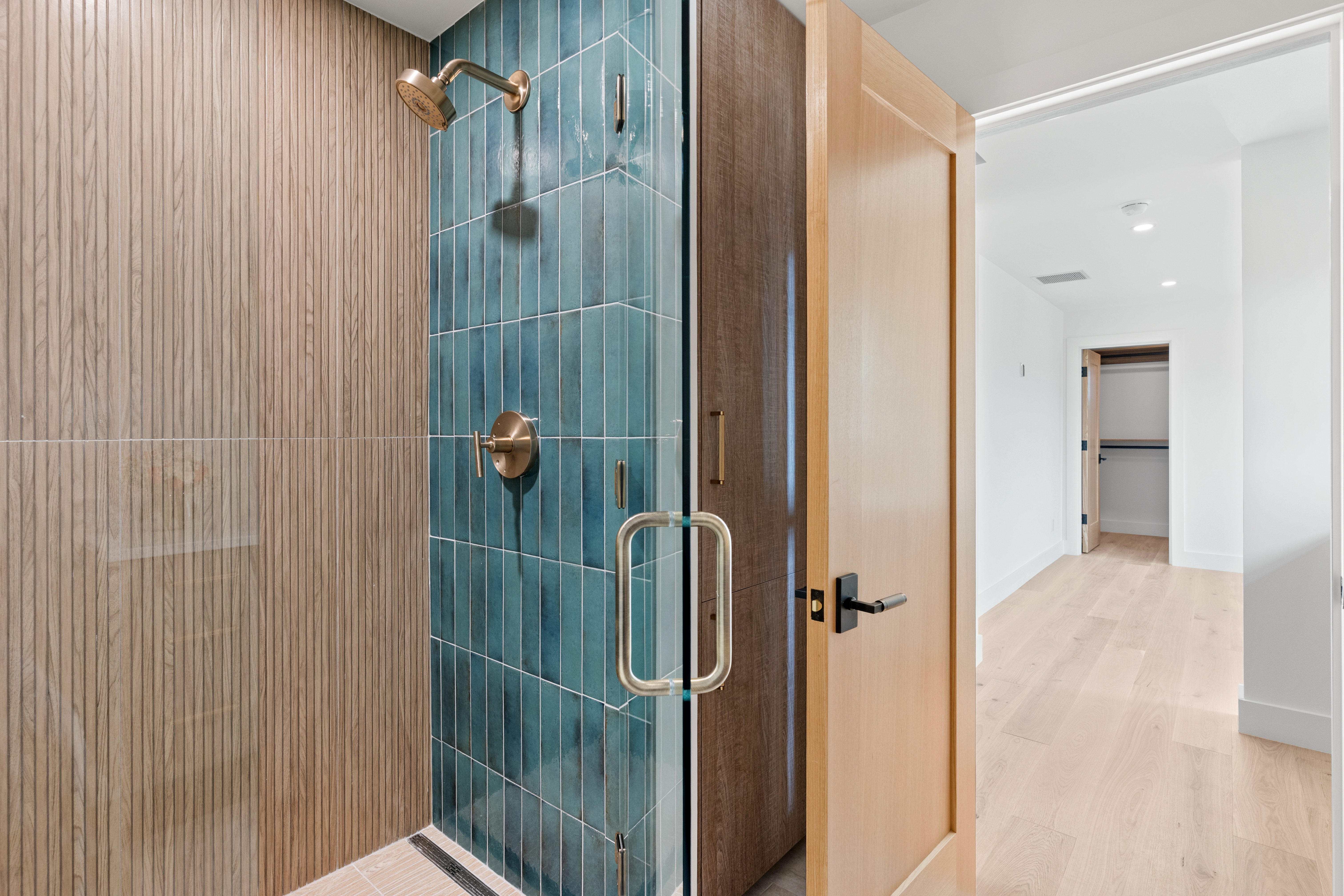 This modern bathroom features a walk-in shower with striking, vertical blue-green subway tiles contrasted by textured, wood-slatted accent walls. The space includes premium brass hardware and a sleek glass enclosure, leading directly into a light-filled hallway with matching hardwood flooring. The overall aesthetic is contemporary, upscale, and sophisticated.