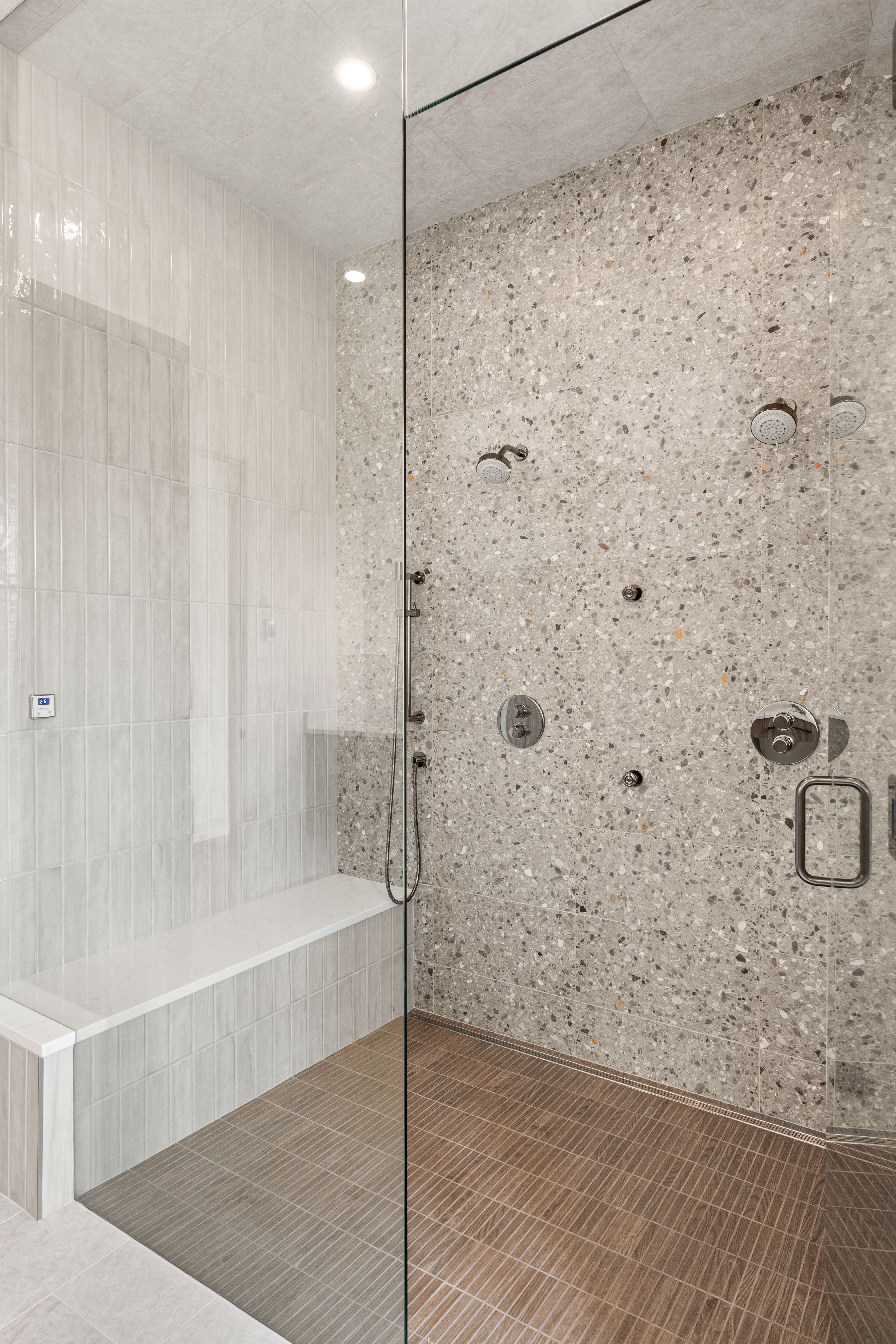 This modern, spa-like walk-in shower features a sophisticated mix of materials, including light-toned vertically stacked tiles on the left and a textured, terrazzo-style stone wall on the right. The space is equipped with a premium multi-head shower system, a built-in bench with a white countertop, and seamless glass paneling, creating an upscale, contemporary aesthetic.