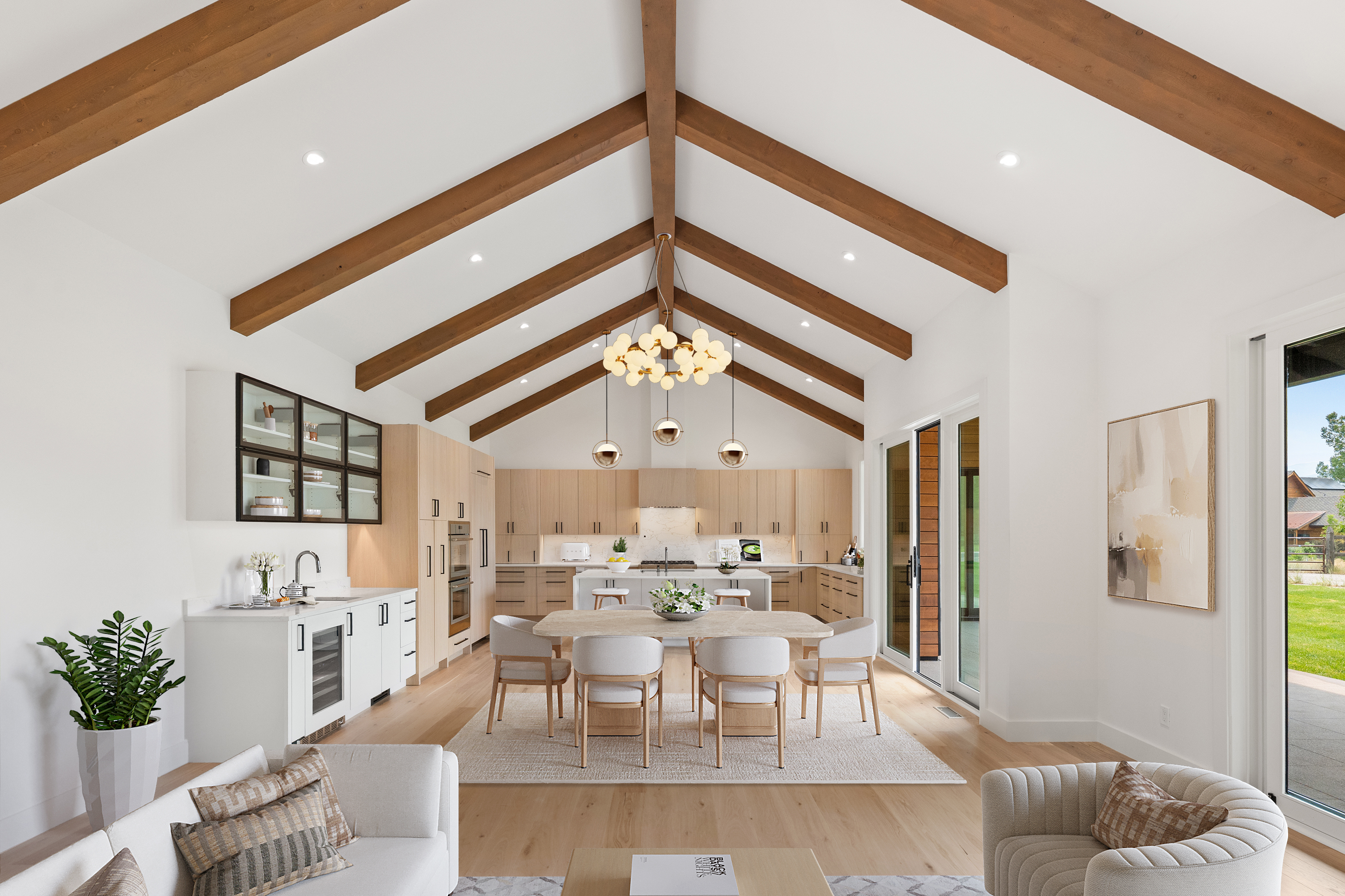 This spacious, open-concept kitchen features a modern aesthetic with light oak cabinetry, a large central island, and soaring vaulted ceilings accented by dramatic wooden beams. The area seamlessly flows into a bright dining space and a lounge, making it ideal for entertaining. High-end finishes, such as a built-in wine cooler and elegant pendant lighting, contribute to a sophisticated and airy atmosphere.