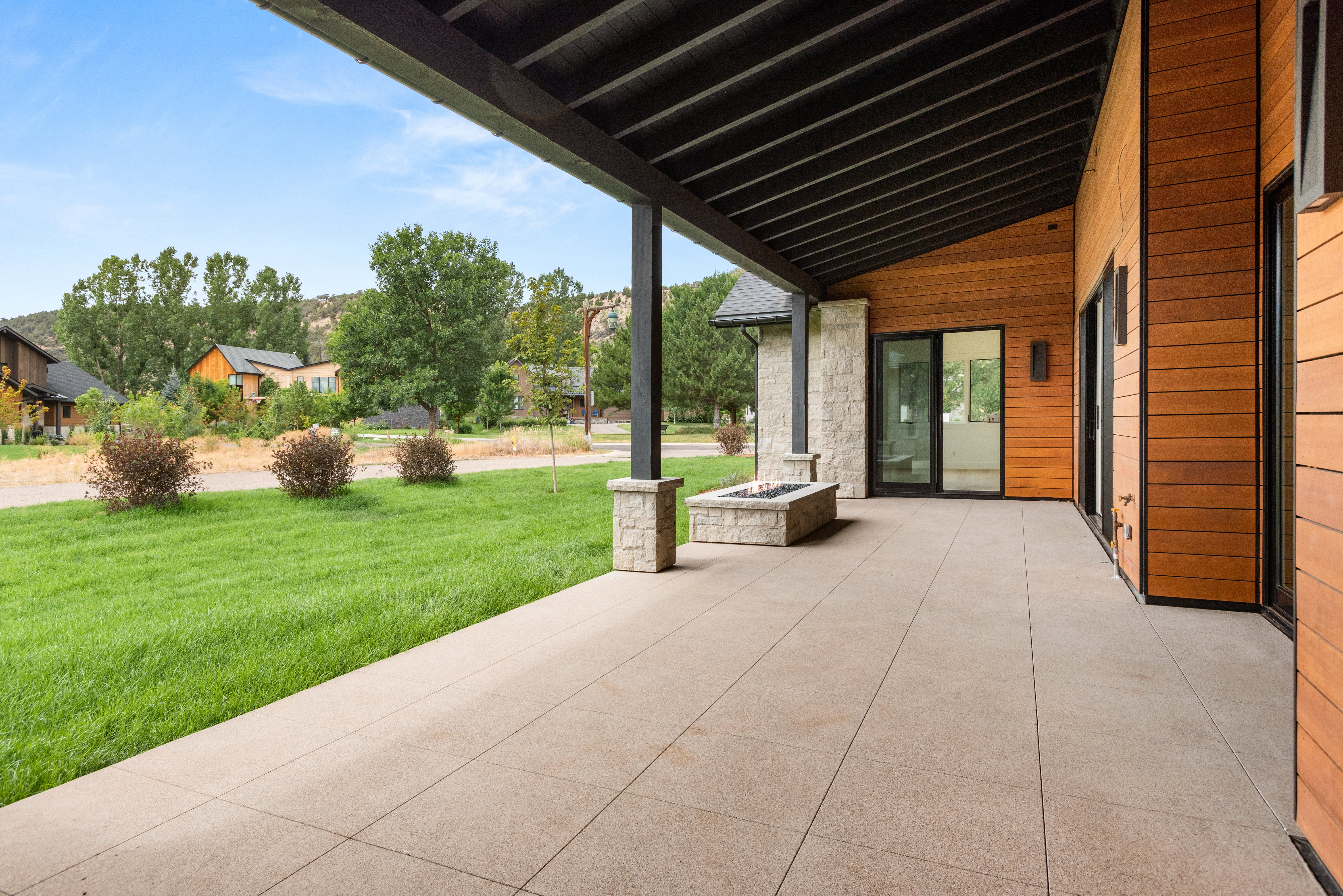 This modern covered patio offers a spacious outdoor living area with large, smooth concrete pavers and a built-in stone fire pit. The space features a contemporary design with warm wood siding, high-contrast dark structural beams, and seamless access to the interior via large sliding glass doors, overlooking a well-manicured lawn and a tranquil neighborhood setting.