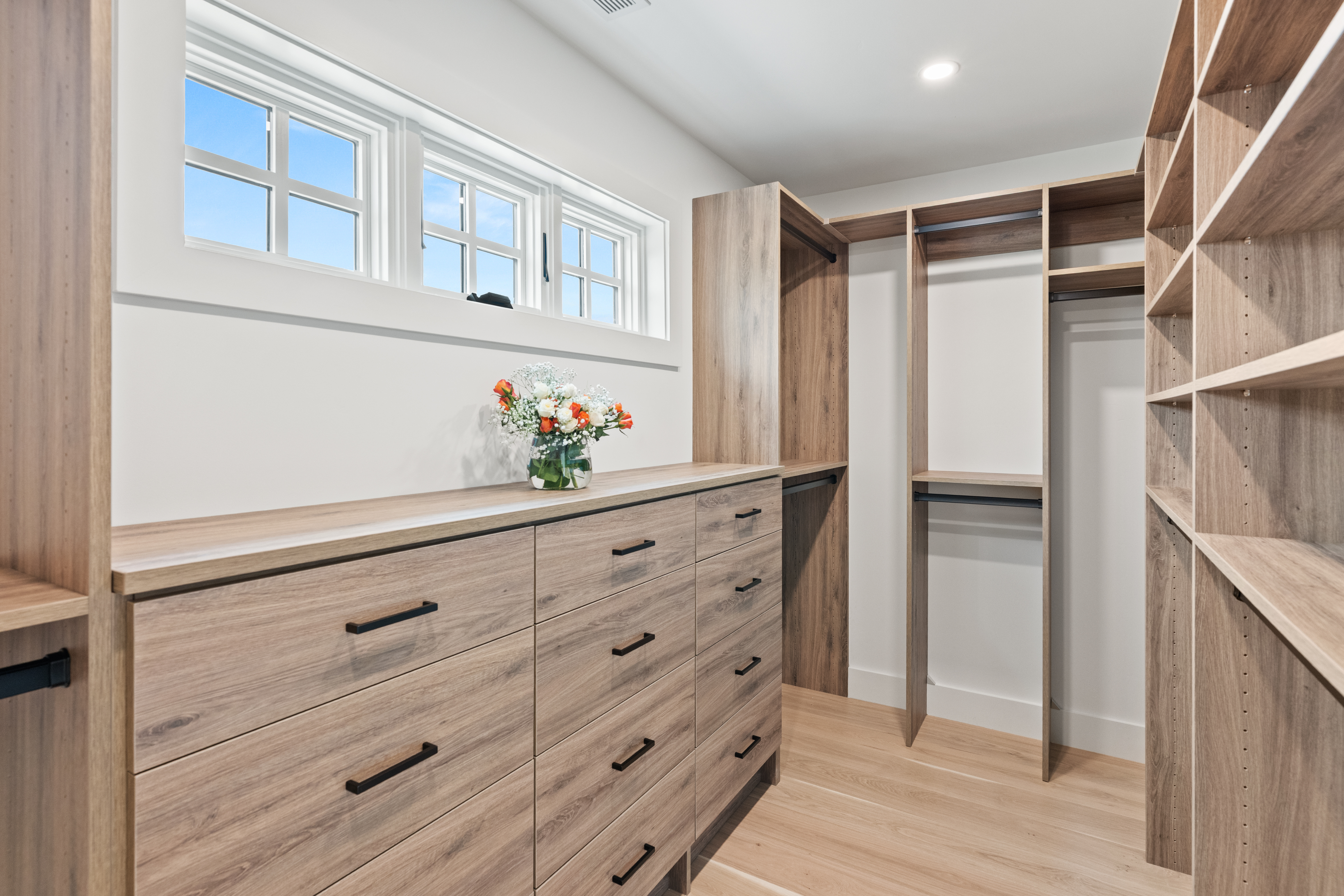 This spacious and organized walk-in closet features contemporary light wood-toned built-in cabinetry, including extensive drawer space and open shelving for storage. A window provides natural light, and the sleek design offers a clean, modern aesthetic perfect for a high-end home. The integration of hanging rods and tailored shelving maximizes functionality while maintaining an upscale, orderly appearance.