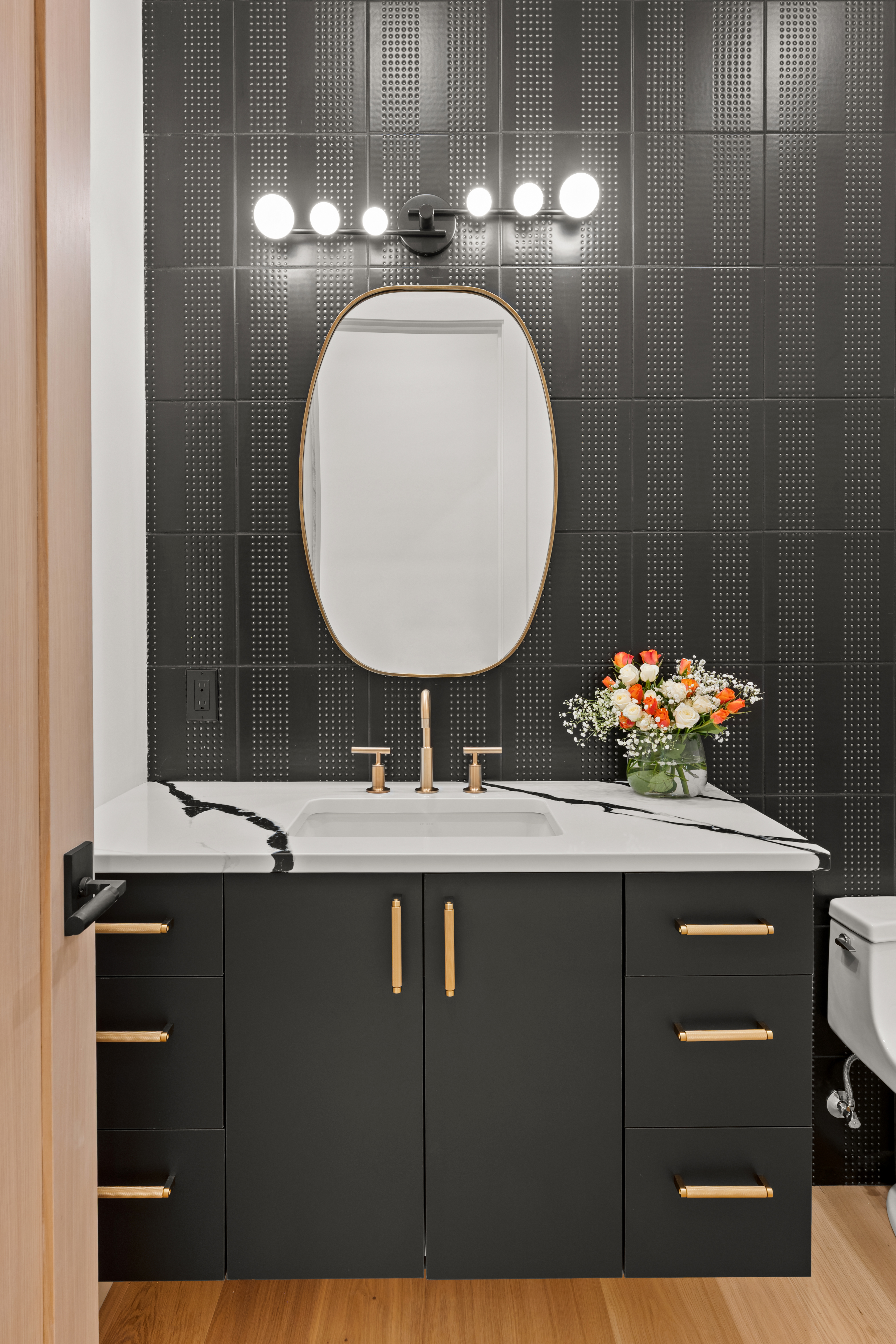 This modern powder room features a sophisticated dark vanity with gold hardware topped by a bold, white marble countertop with black veining. The wall is accented by black textured tiles that create a striking backdrop for the sleek, oval-shaped mirror and minimalist vanity light fixture. A vase of fresh flowers adds a touch of elegance to the high-contrast, contemporary space.