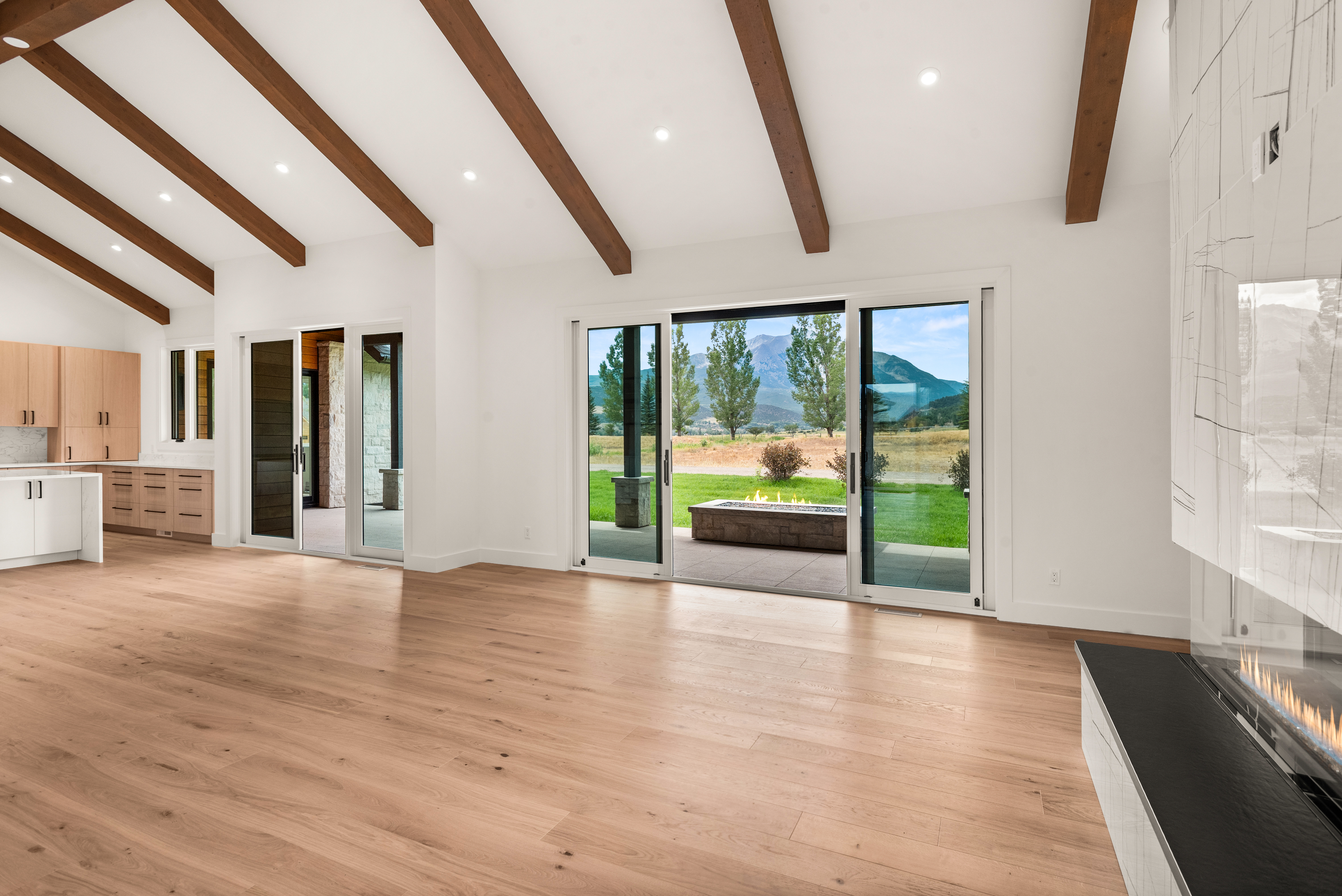 This spacious, modern living area features vaulted ceilings with exposed wood beams and light-toned hardwood flooring throughout. Large sliding glass doors open to a patio with a built-in fire pit and scenic mountain views, seamlessly blending indoor and outdoor living. The space is highlighted by a sleek, modern fireplace on the right and an adjacent open-concept kitchen with light wood cabinetry.