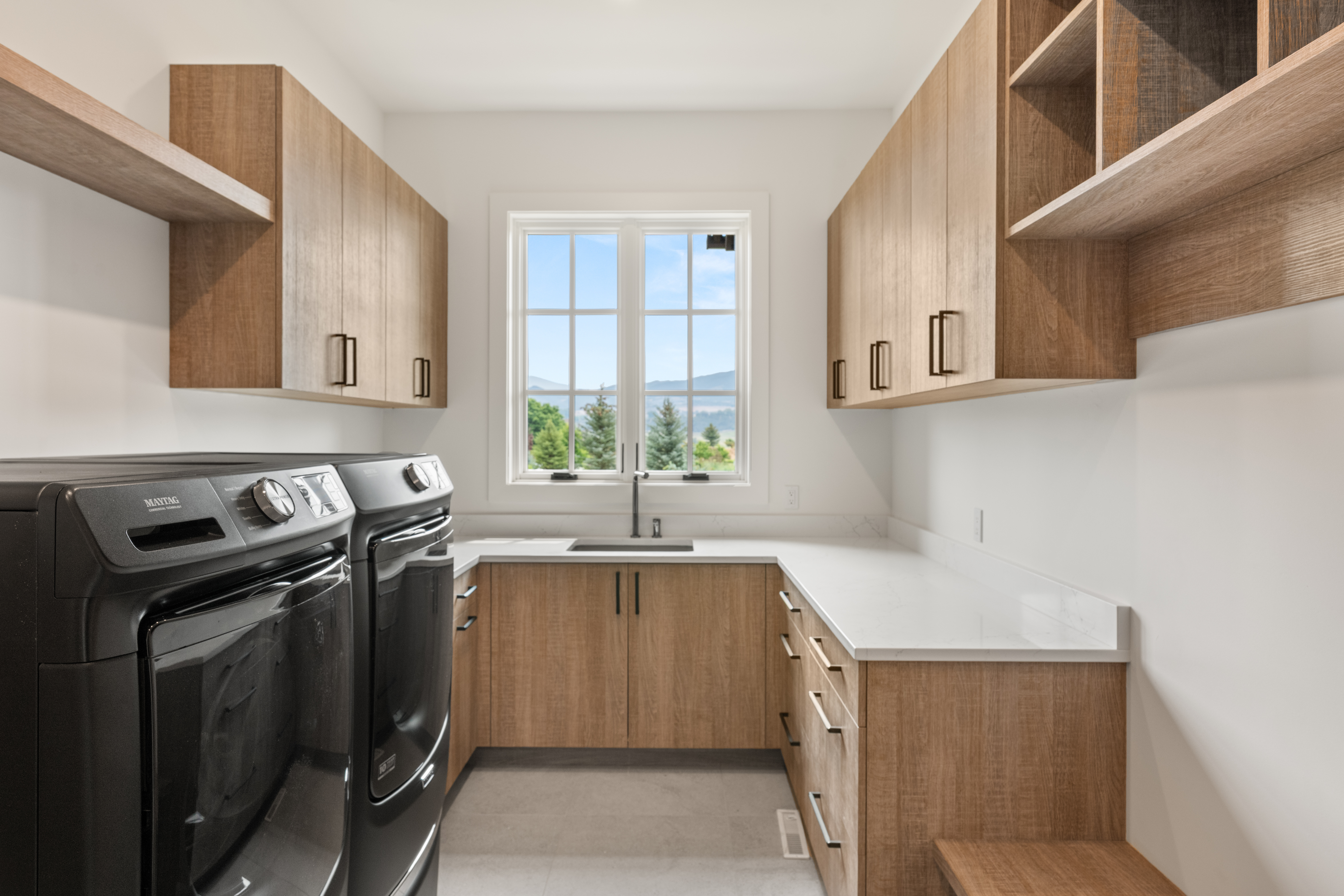 This clean and functional laundry room features warm wood-toned cabinetry, bright white countertops, and sleek black hardware. The room includes side-by-side Maytag laundry appliances and a built-in sink located directly beneath a large window that offers natural light and scenic views. The combination of light walls and natural wood provides a modern, inviting aesthetic suitable for a high-end home.