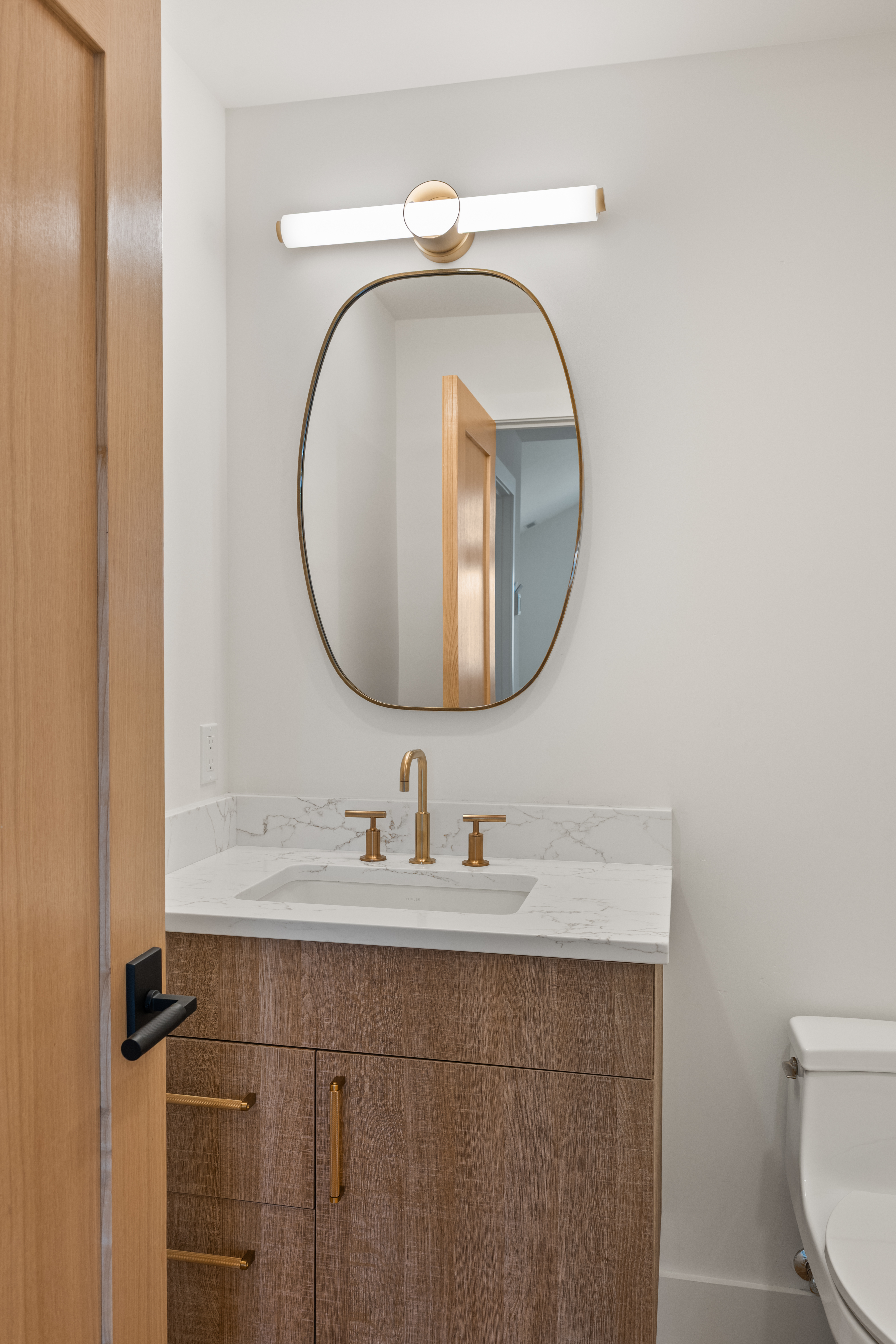 This modern powder room features a sleek vanity with a wood-grain finish, topped by a white marble-look countertop and a gold-toned faucet. The room includes a contemporary oval-shaped wall mirror and a matching minimalist light fixture, creating a sophisticated and clean aesthetic. A portion of the toilet is visible on the right, and the space is accessed through a natural wood-toned door with a bold black handle.