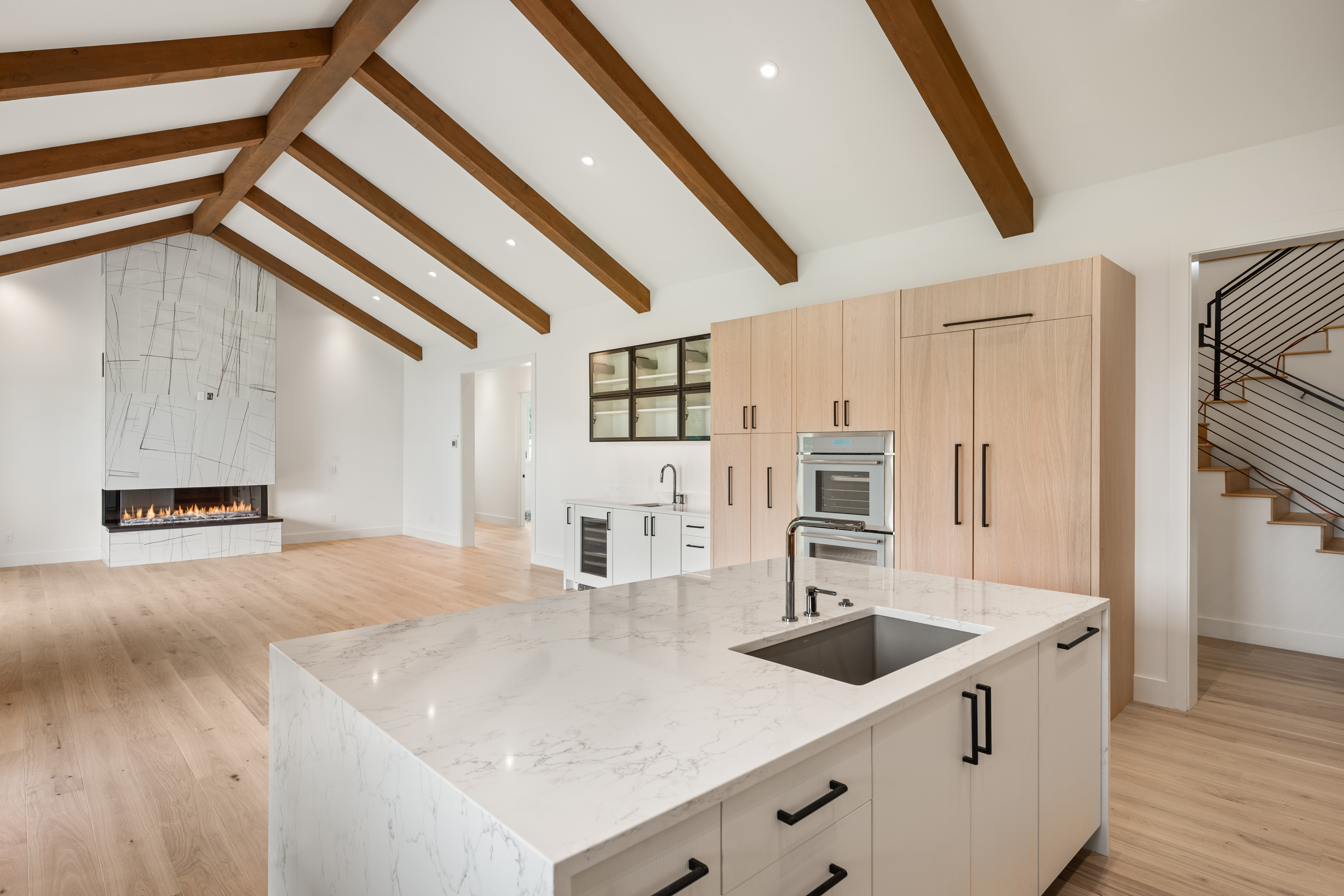 This stunning open-concept kitchen features a modern aesthetic with light oak wood cabinetry, sleek matte black hardware, and a waterfall-edge marble-look island. Vaulted ceilings with exposed wooden beams add architectural character, while a statement fireplace with marble cladding anchors the adjacent living space. The bright, neutral palette and high-end finishes create a sophisticated and airy atmosphere perfect for modern living.