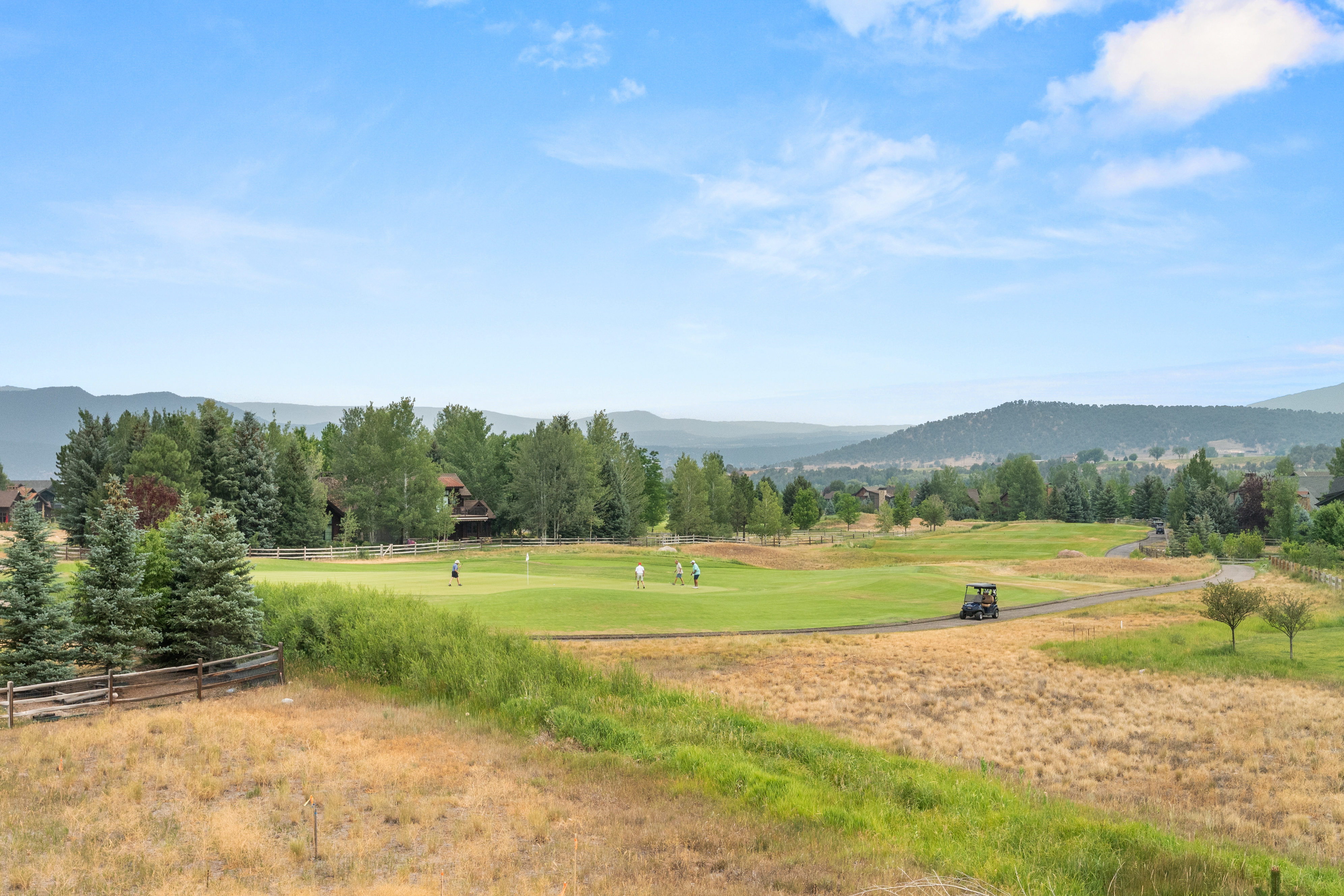 This image showcases a scenic golf course fairway with a well-manicured putting green and players enjoying a round. Surrounded by lush trees, mountain vistas, and charming residential homes, the setting provides a serene and inviting community atmosphere. The presence of a golf cart path and natural landscape highlights the recreational appeal of this residential neighborhood.