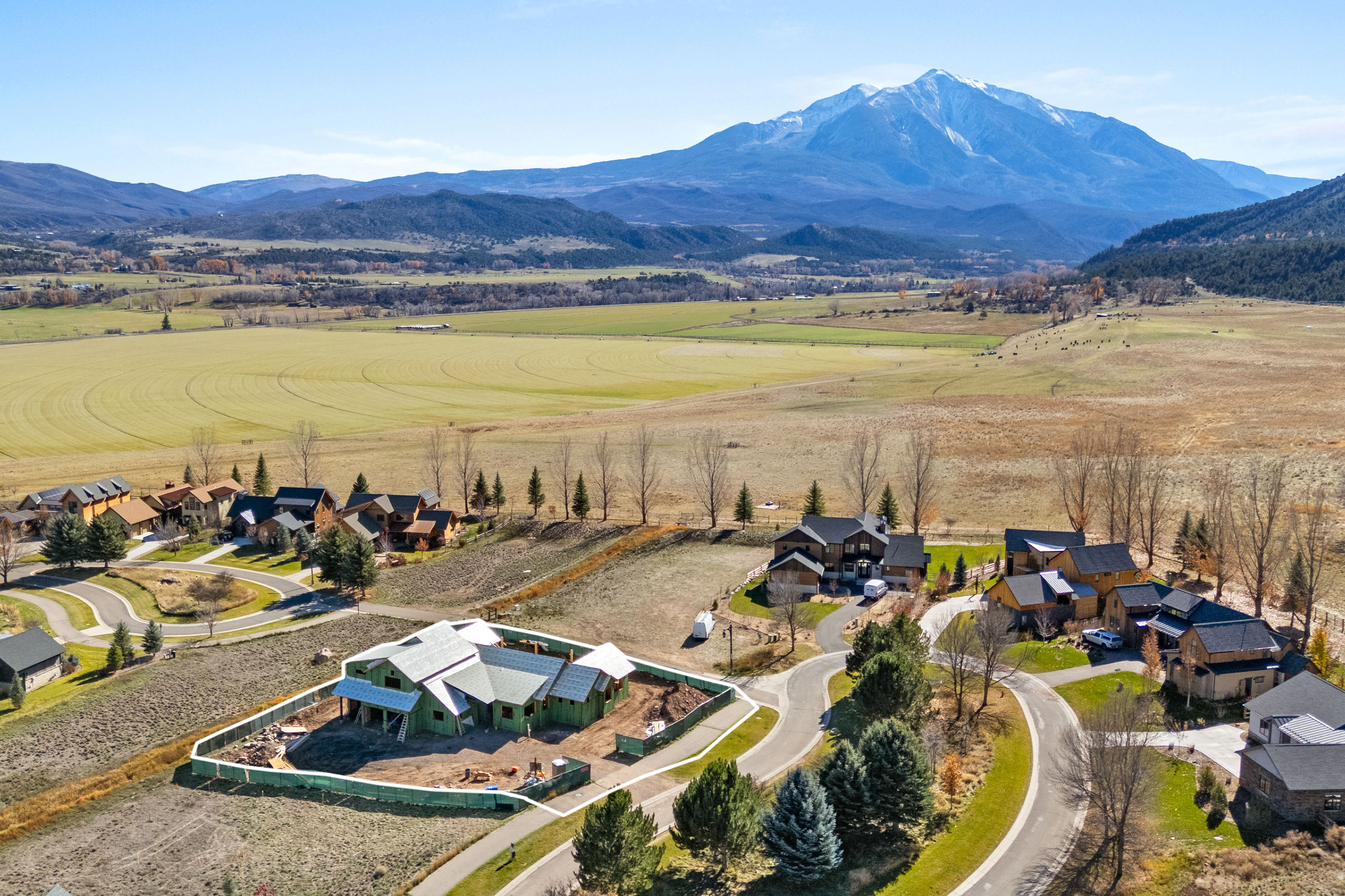 This aerial view captures a picturesque residential development set against a stunning, snow-capped mountain backdrop. The landscape features a mix of completed luxury homes and a single residence currently under construction, surrounded by expansive, open fields and natural vegetation. The serene setting and well-planned community layout offer an inviting atmosphere, emphasizing mountain living with convenient access to nature.
