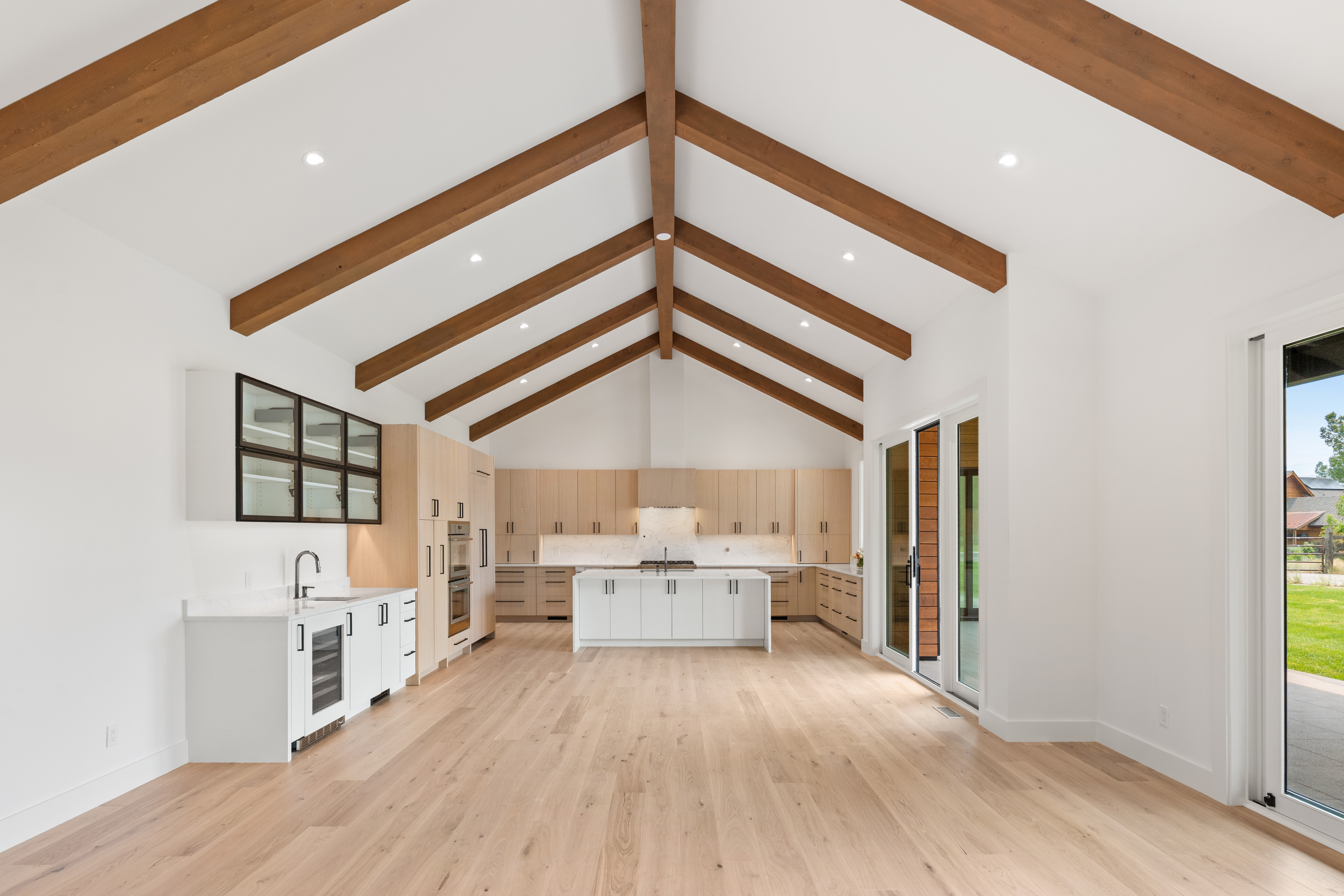 This stunning open-concept kitchen features a dramatic vaulted ceiling with exposed wooden beams and high-end natural wood cabinetry. The space is anchored by a large central island with quartz countertops and includes professional-grade appliances, clean white accents, and expansive sliding glass doors that lead to the outdoors. The bright, airy design and modern finishes make this an exceptional space for both cooking and entertaining.