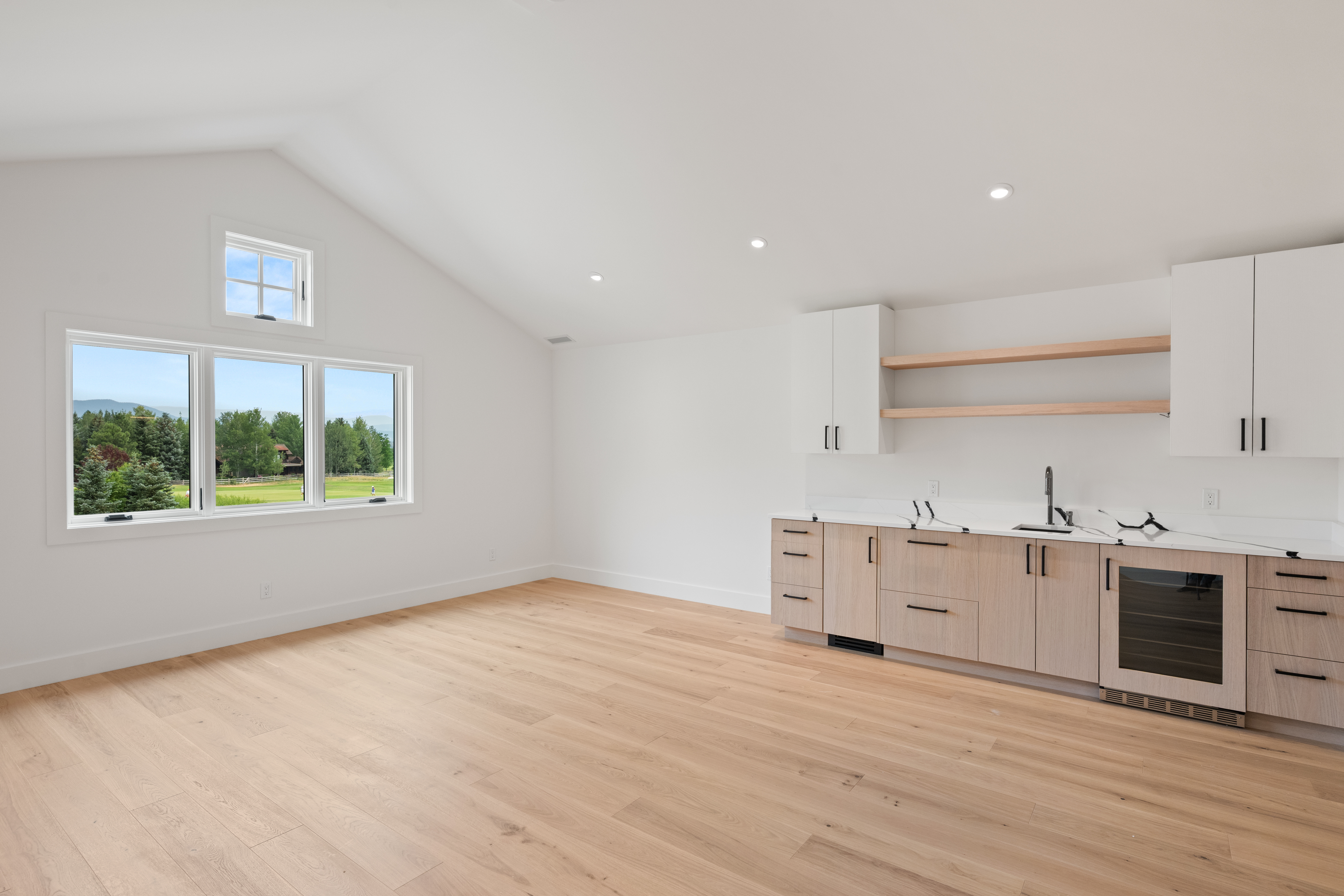 This bright and airy kitchenette features a contemporary design with light wood cabinetry, quartz countertops, and floating shelves. The vaulted ceiling and expansive windows fill the space with natural light while offering scenic outdoor views. It is a stylish and functional wet bar or auxiliary kitchen area suitable for a modern home.