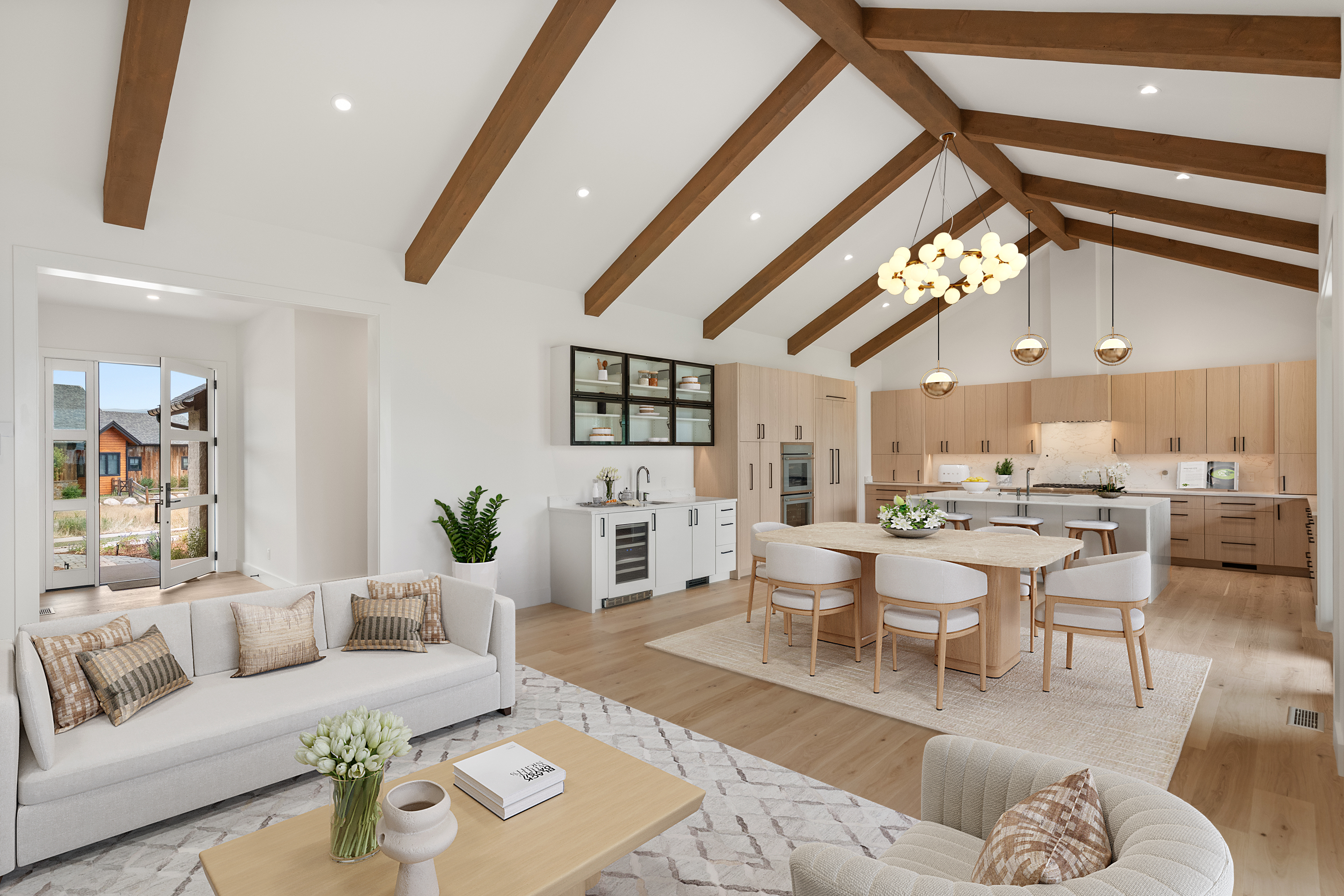 This spacious, open-concept living area seamlessly integrates a modern living room, dining space, and gourmet kitchen under vaulted ceilings with exposed wood beams. The room features high-end finishes, light wood flooring, and a neutral color palette that emphasizes natural light, creating a bright and airy atmosphere. The inclusion of a dedicated wet bar and direct access to an outdoor entryway adds both functionality and a welcoming, luxurious feel.