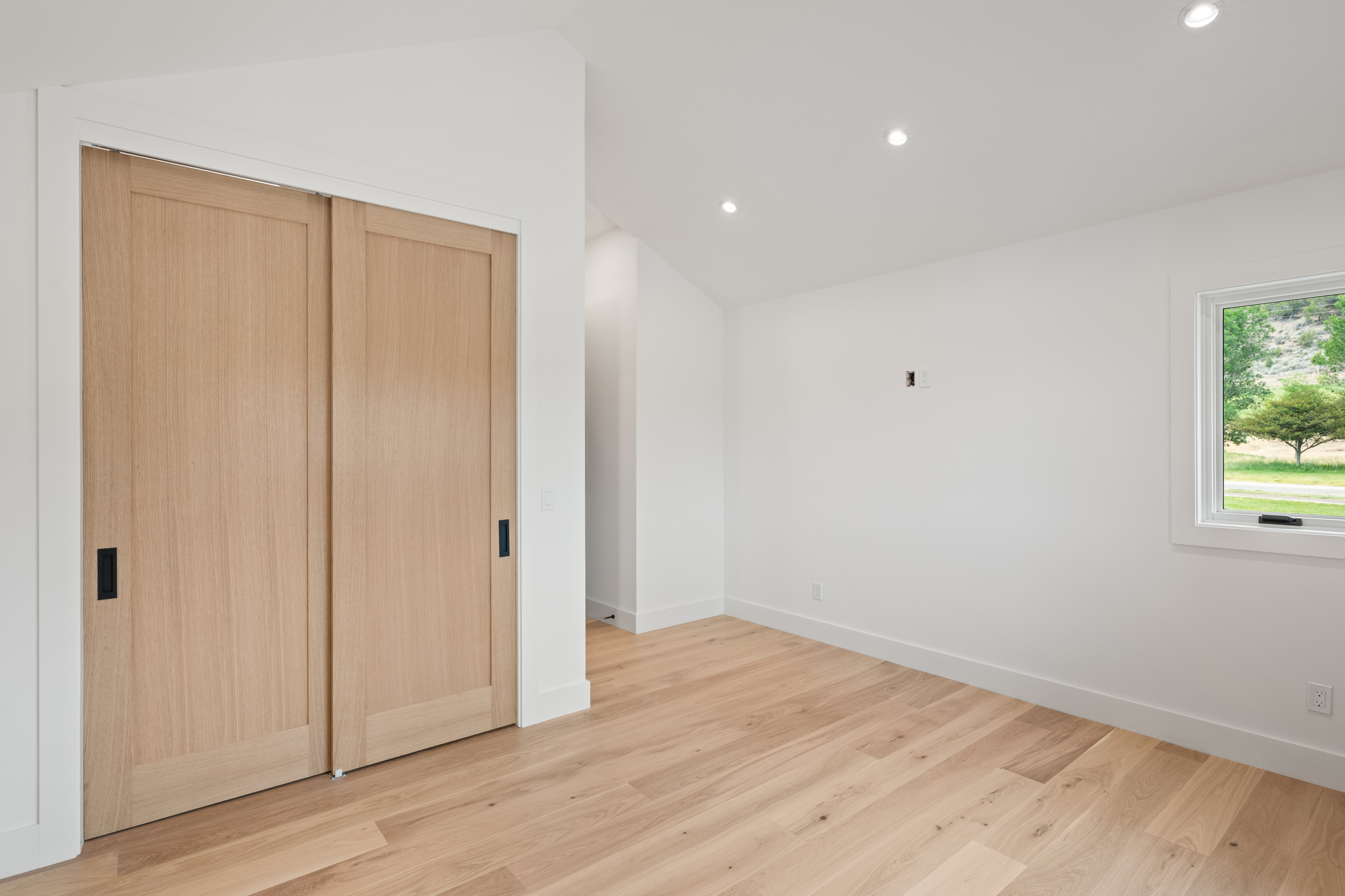 This clean, minimalist room features light hardwood flooring and neutral white walls, offering a bright and airy atmosphere. The space includes a pair of sliding wooden closet doors, recessed lighting, and a window with a pleasant view of the natural surroundings. It presents a versatile, modern aesthetic perfect for a bedroom, office, or creative space.