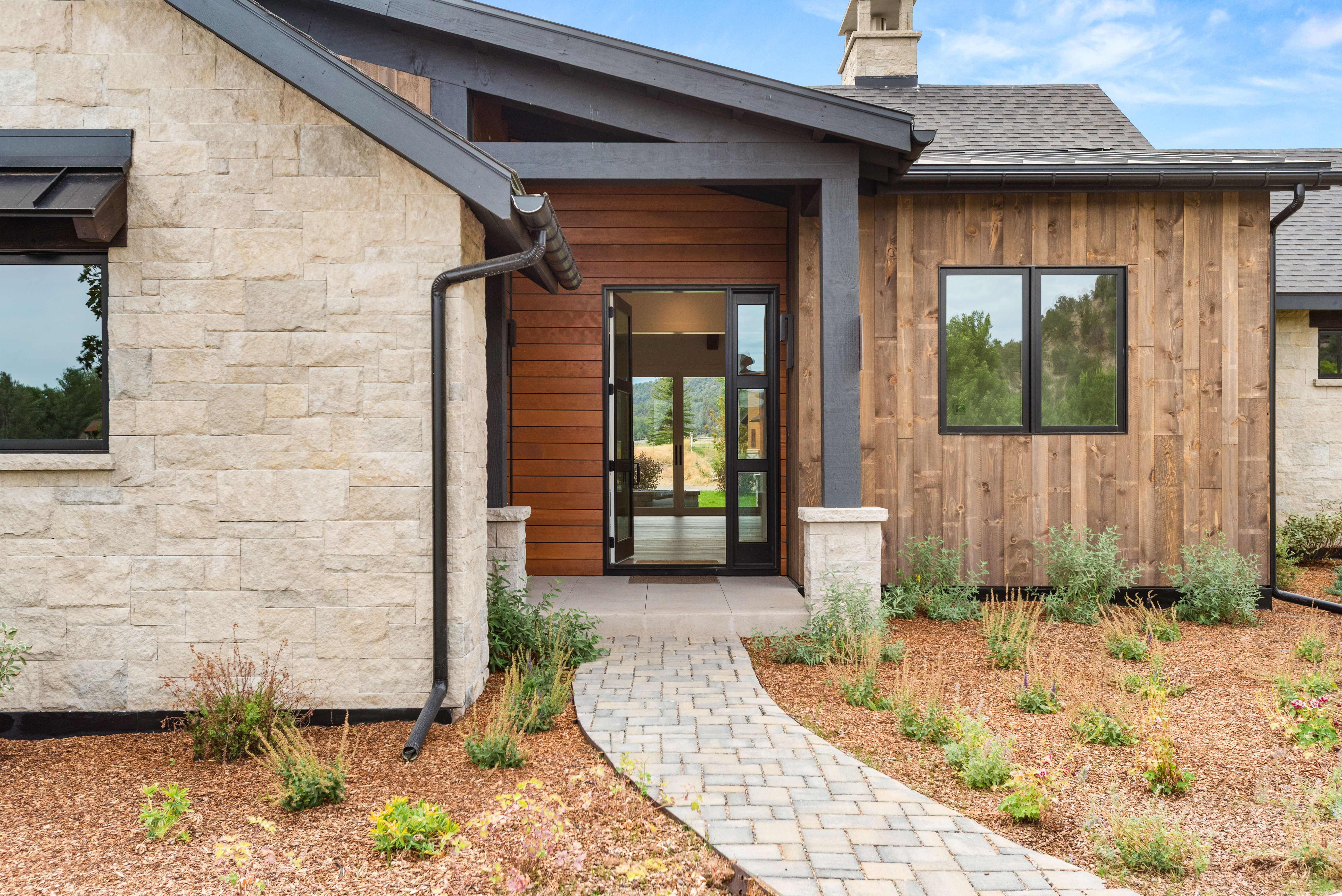 This inviting entryway showcases a blend of modern design with natural textures, featuring stacked stone siding paired with warm wood accents. The path leads to a sleek, dark-framed glass door, while well-maintained landscaping and mulch add to the home's curb appeal. The overall aesthetic is sophisticated, rustic, and welcoming.