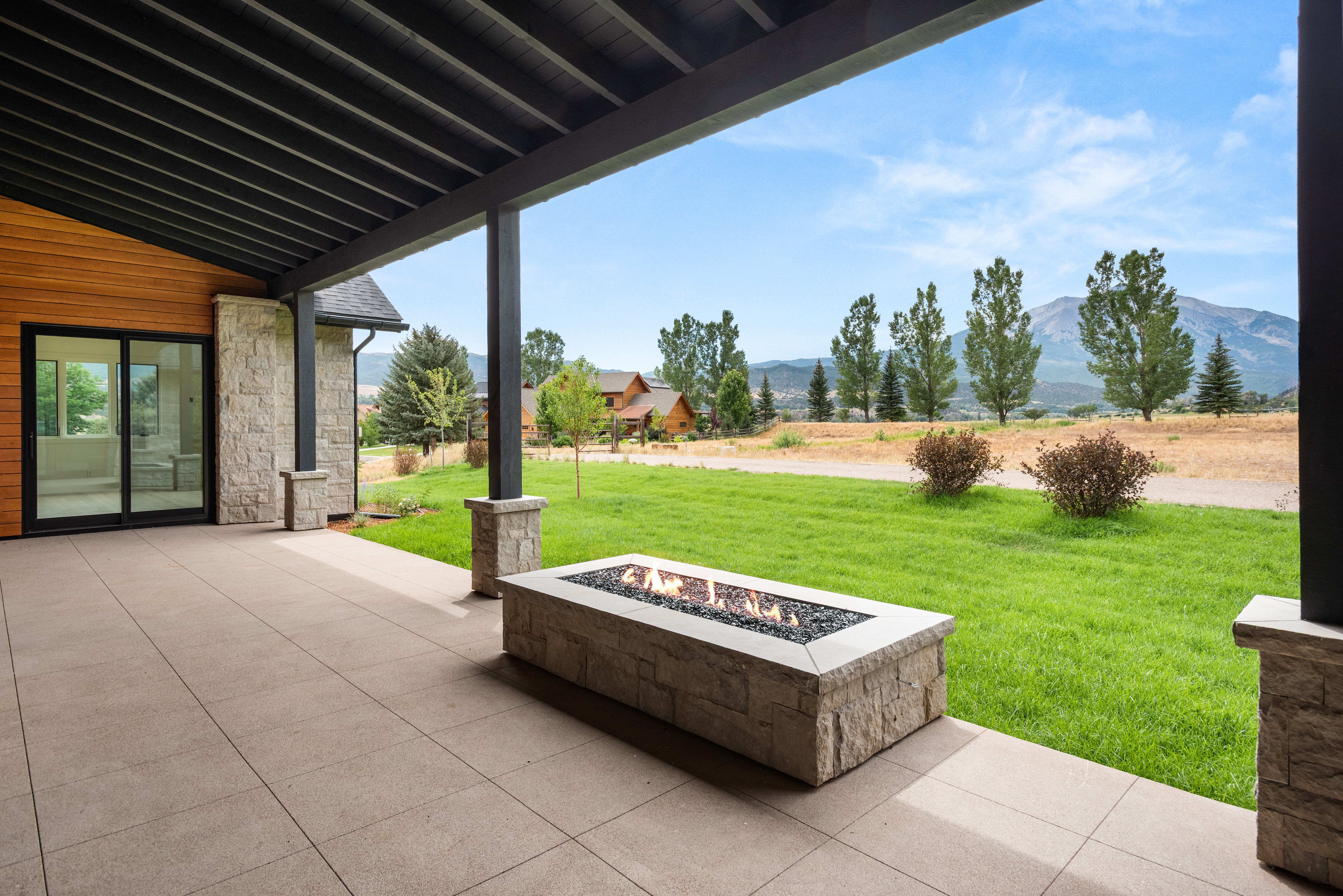 This covered patio space offers a seamless transition between indoor and outdoor living, featuring a sleek, rectangular stone fire pit as a central gathering point. The expansive area is floored with large, neutral-toned pavers and is framed by stone-accented columns under a wooden ceiling structure. Beyond the patio, a well-manicured lawn meets a picturesque rural landscape with mature trees and mountain views, highlighting the property's serene and spacious setting.