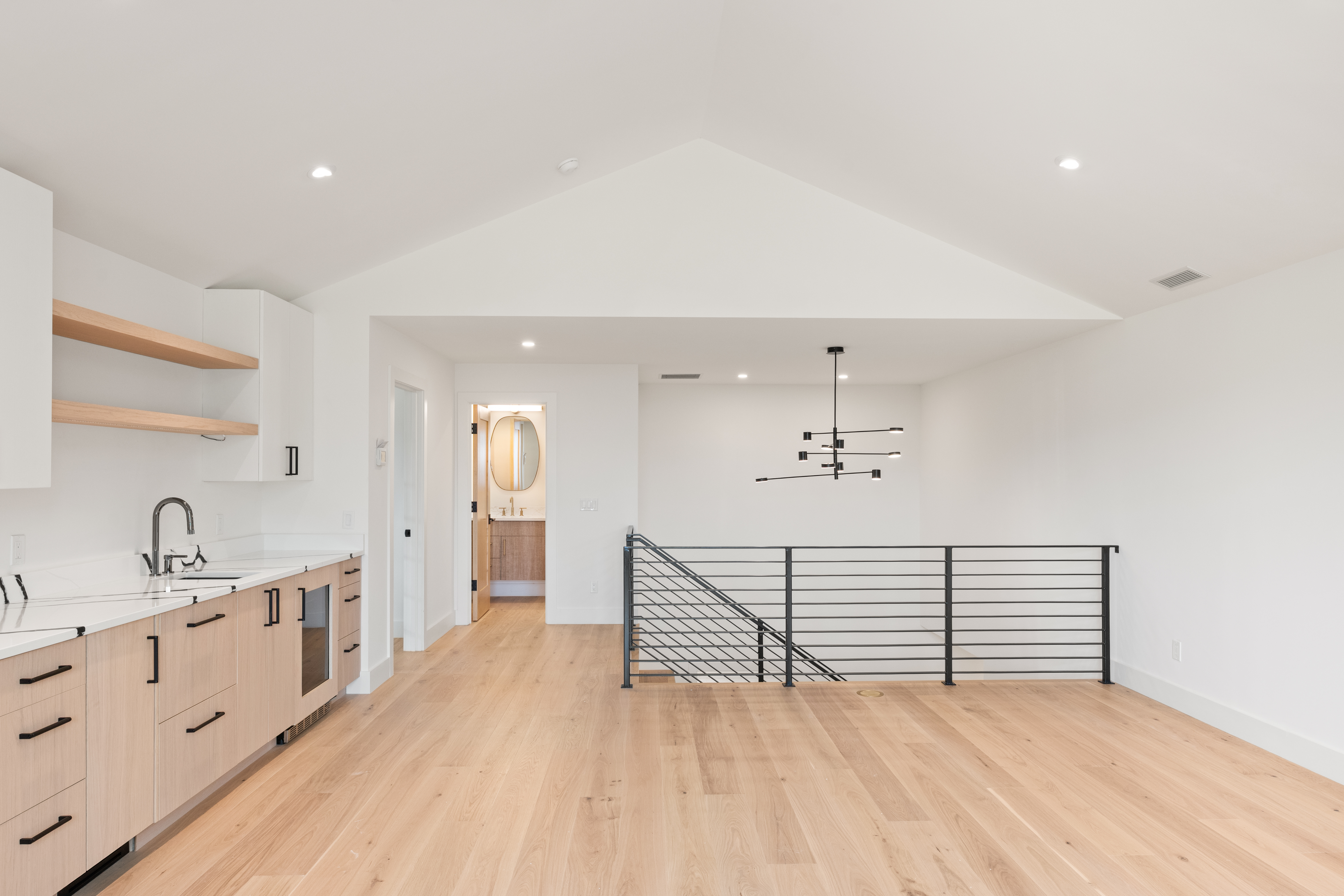 This bright, contemporary kitchenette features light wood cabinetry, white countertops, and open shelving, creating a clean and minimalist aesthetic. The space is open and airy, featuring vaulted ceilings and a sleek black metal railing overlooking the staircase. The inclusion of an under-counter beverage fridge and proximity to a bathroom suggests a versatile functional space suitable for a loft, wet bar, or entertainment area.