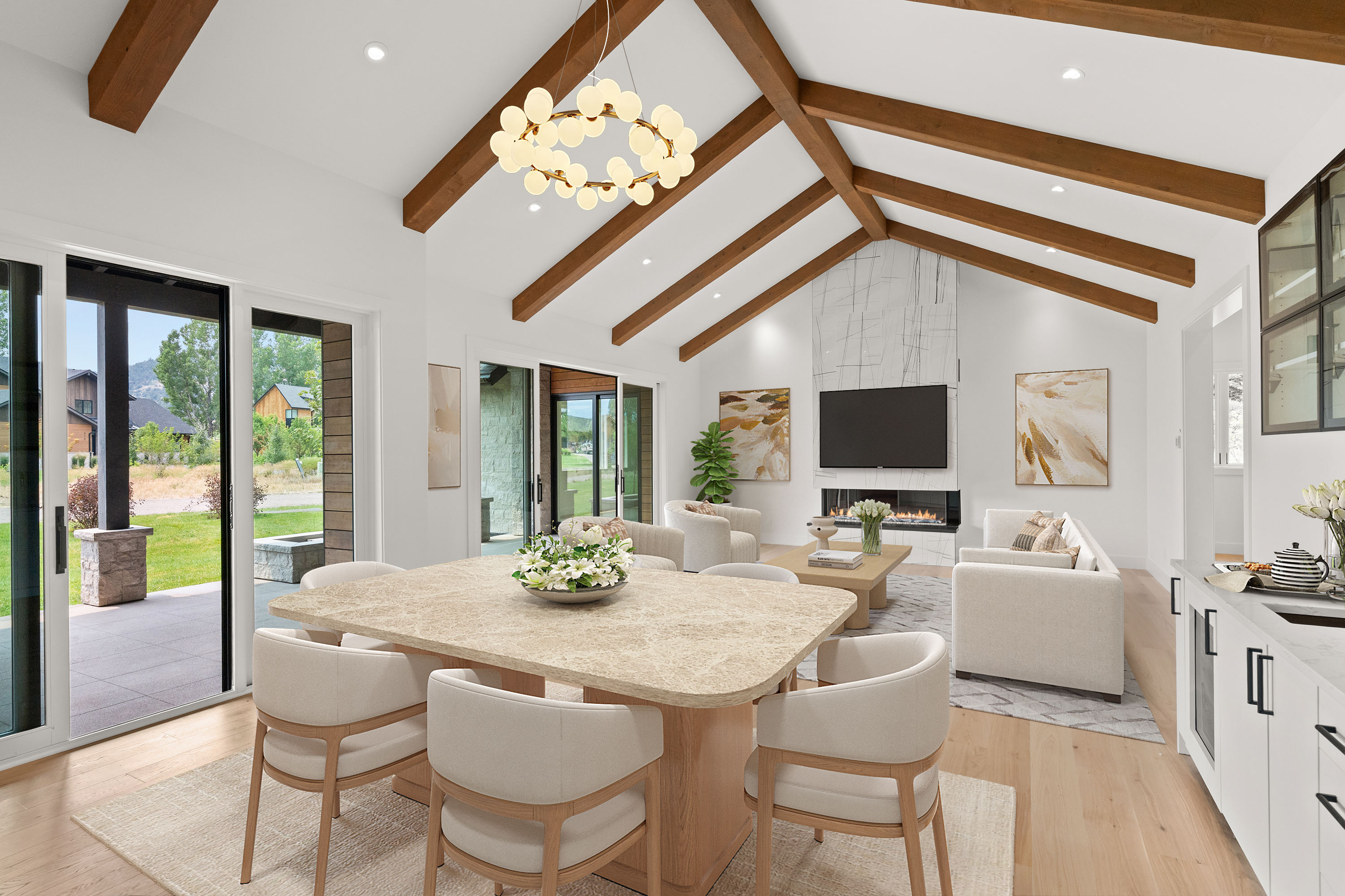 This spacious, open-concept living and dining area features vaulted ceilings with exposed wood beams and a stunning floor-to-ceiling marble fireplace. The design emphasizes natural light with expansive sliding glass doors that provide seamless indoor-outdoor living, complemented by sophisticated, neutral furnishings. It is a luxurious, modern space perfect for entertaining.