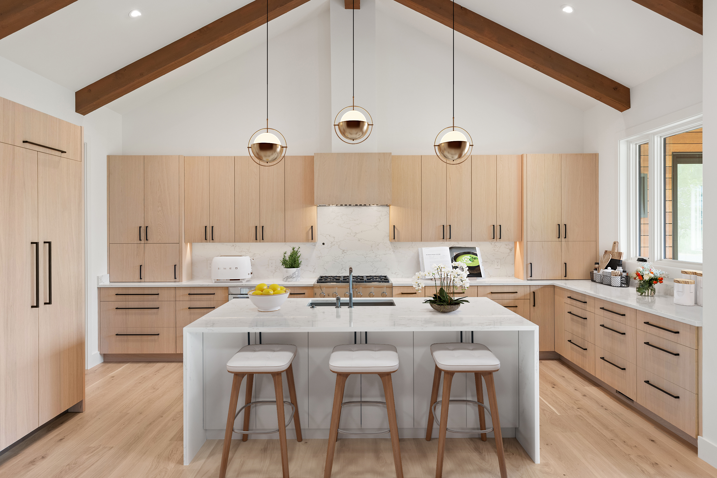 This stunning modern kitchen features warm light-wood cabinetry paired with sleek white marble countertops and a matching central island. The vaulted ceiling with exposed wooden beams adds an architectural flair, while three brass globe pendant lights provide elegant task lighting over the island. A high-end gas range with a custom hood and an integrated farmhouse sink complete this bright, high-end culinary space.