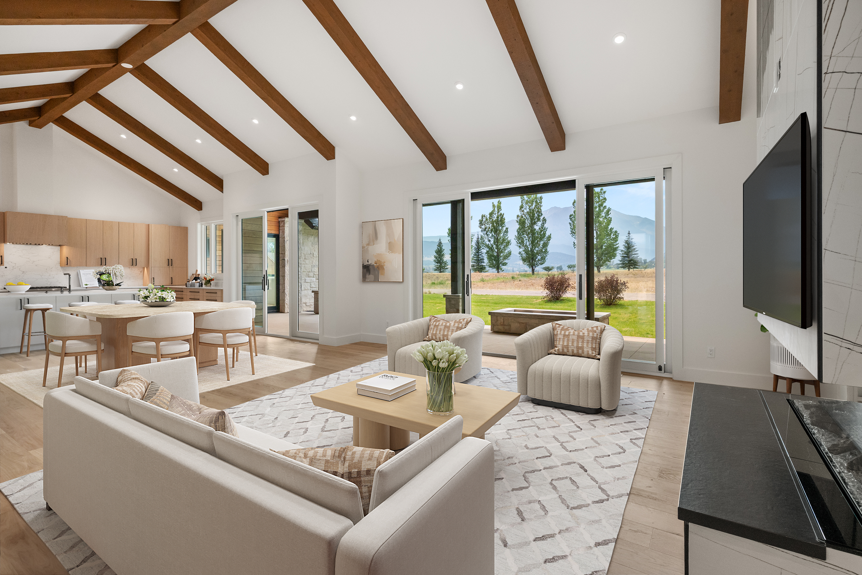 This spacious, open-concept living area features a dramatic vaulted ceiling with exposed wooden beams and large glass sliding doors that reveal a picturesque mountain view. The room is decorated in a sophisticated, neutral palette with contemporary furniture, a textured area rug, and a sleek integrated television, creating an inviting space perfect for entertaining. The seamless connection to the adjacent dining area and kitchen enhances the home's airy, light-filled atmosphere.