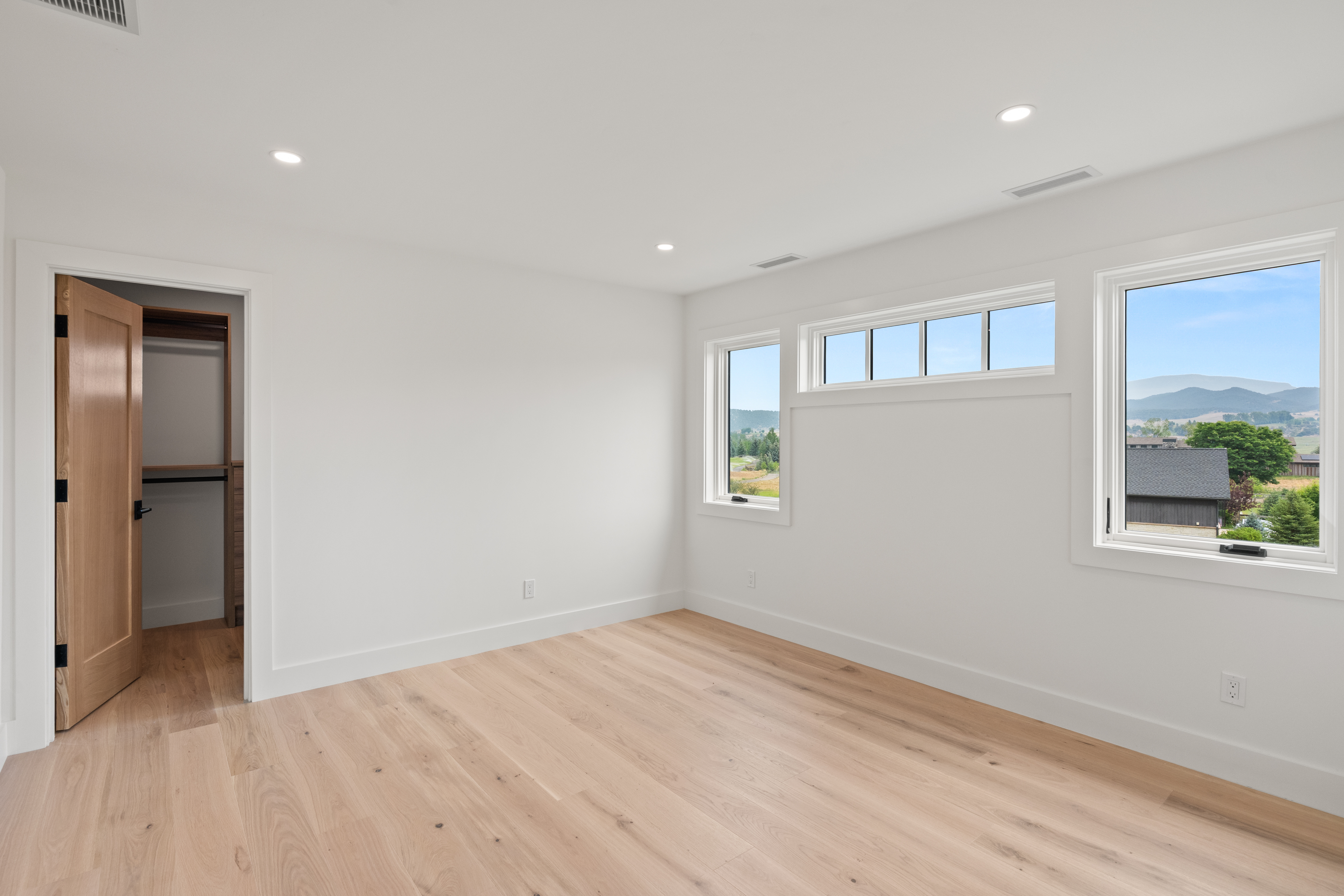 This modern bedroom features clean white walls, light wood flooring, and recessed ceiling lighting, creating a bright and airy atmosphere. The room includes a functional reach-in closet with natural wood accents and large windows that offer scenic outdoor views. It presents a minimalist, contemporary aesthetic that serves as a versatile blank canvas for any decor style.