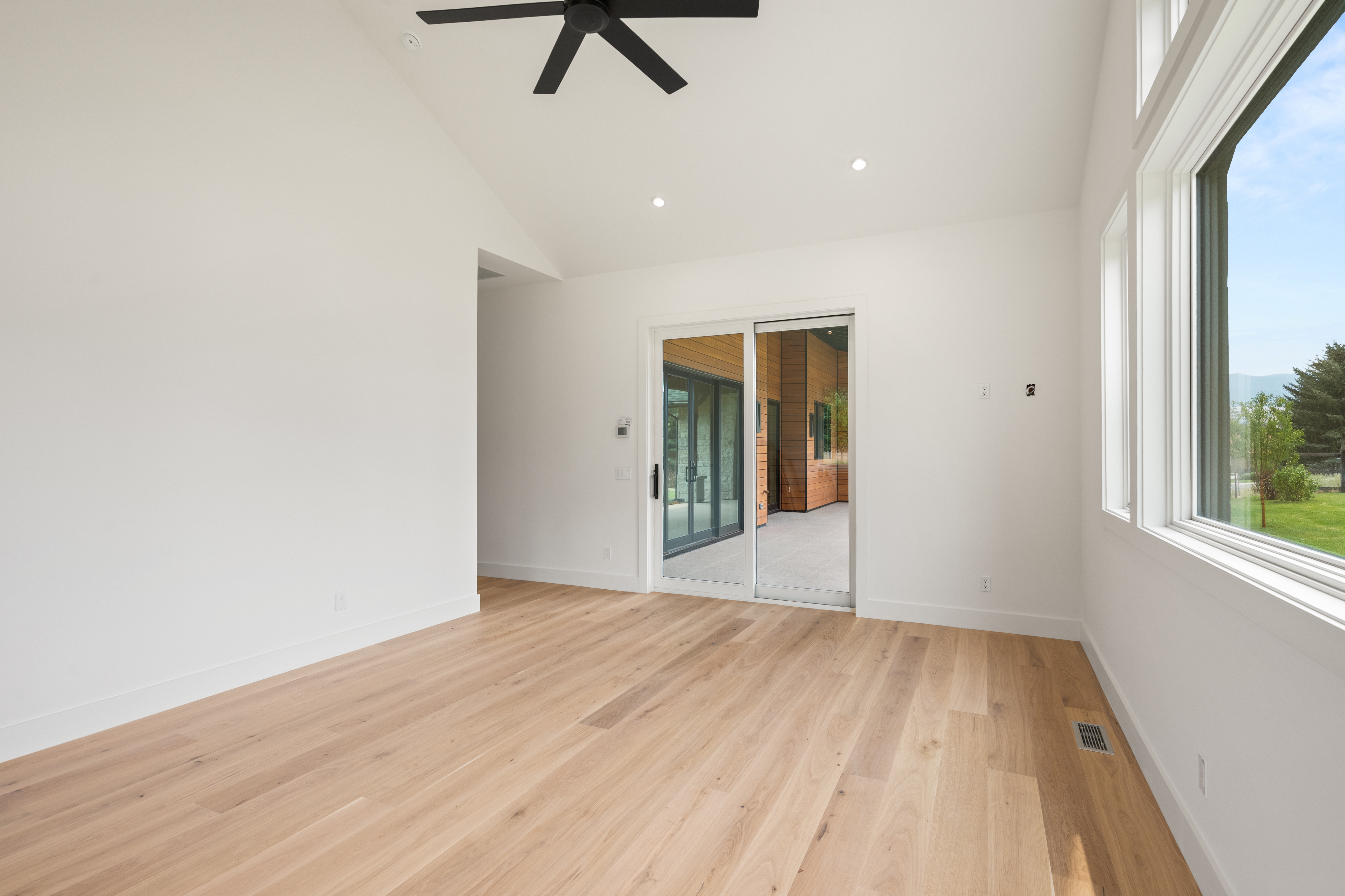 This spacious, light-filled living area features clean white walls and warm, wide-plank hardwood flooring that creates an inviting atmosphere. A large, vaulted ceiling with a modern black ceiling fan adds architectural character, while a sliding glass door provides a seamless transition to the outdoor patio. Large windows on the right allow for ample natural light and offer peaceful views of the surrounding greenery.