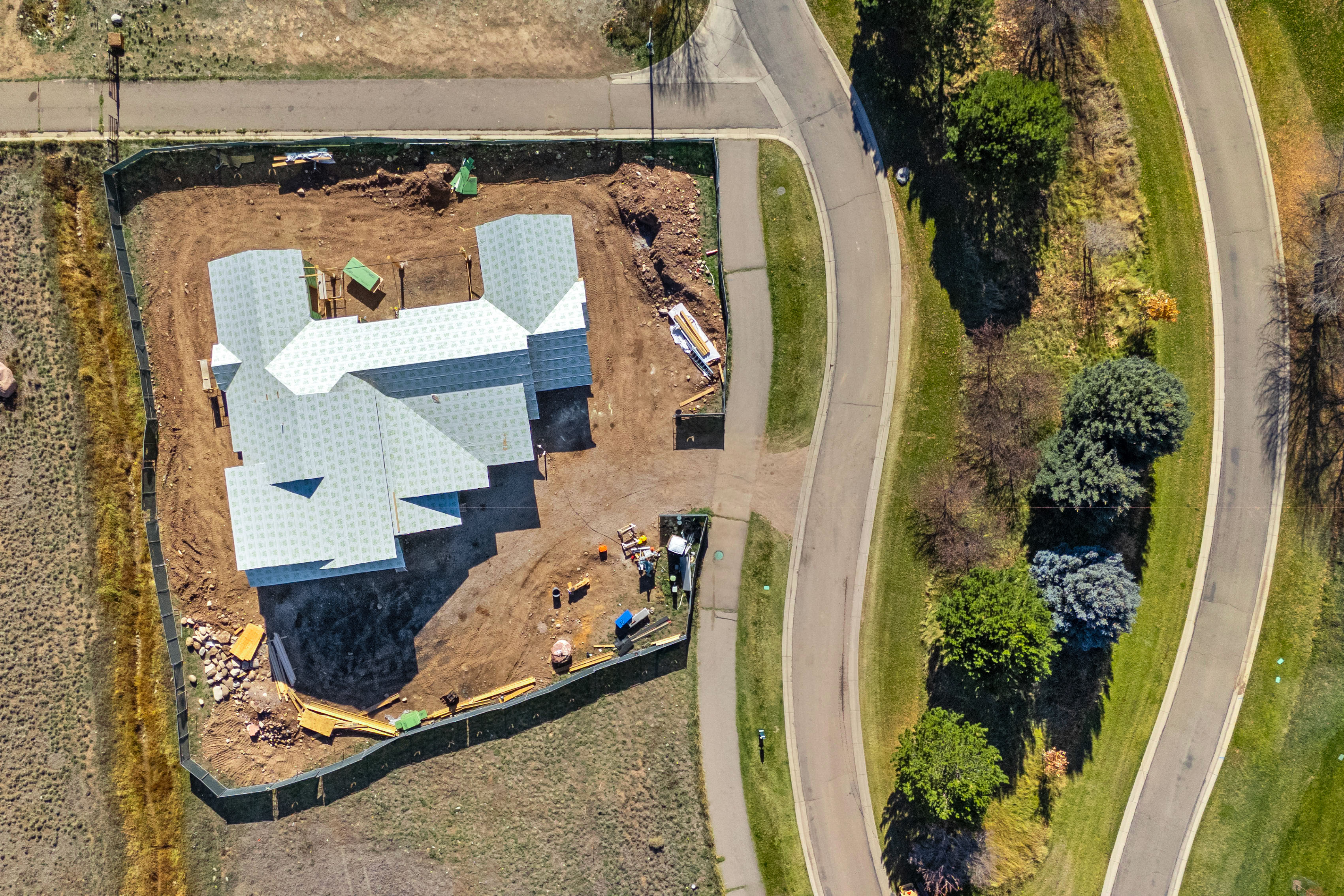 This aerial view showcases a residential construction site in a newly developed neighborhood with a curved road and landscaping. The property features a large, complex roof structure partially completed with sheathing, situated on a generous lot enclosed by a construction fence. The surrounding area includes established trees and green spaces, highlighting the potential for a suburban retreat once the home is finalized.