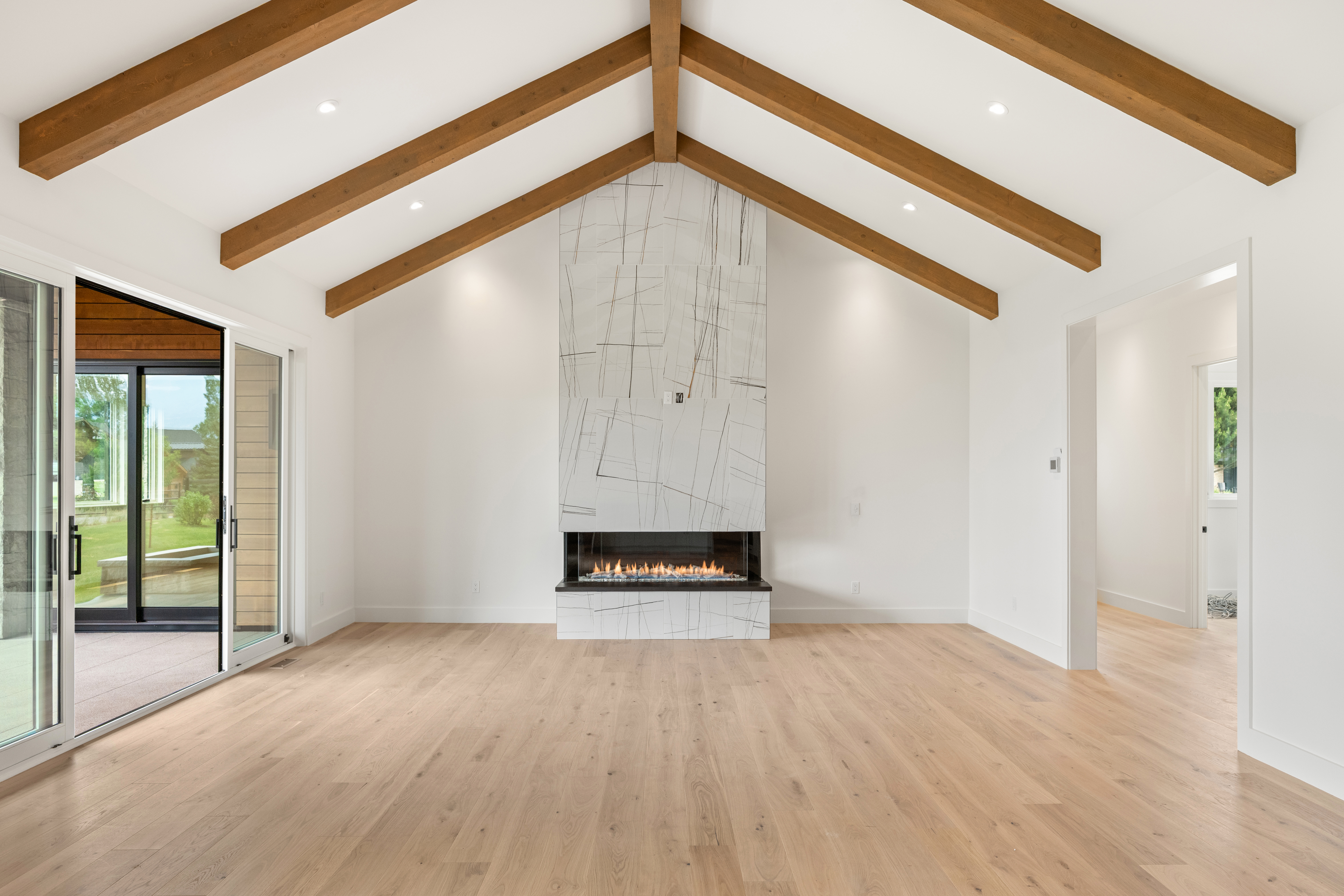This spacious living room features high vaulted ceilings with exposed wood beams and light-colored hardwood flooring, creating an airy and modern aesthetic. The focal point of the room is a striking floor-to-ceiling fireplace clad in white marble tile with charcoal veining. Large sliding glass doors open onto a patio, allowing for seamless indoor-outdoor living and ample natural light.