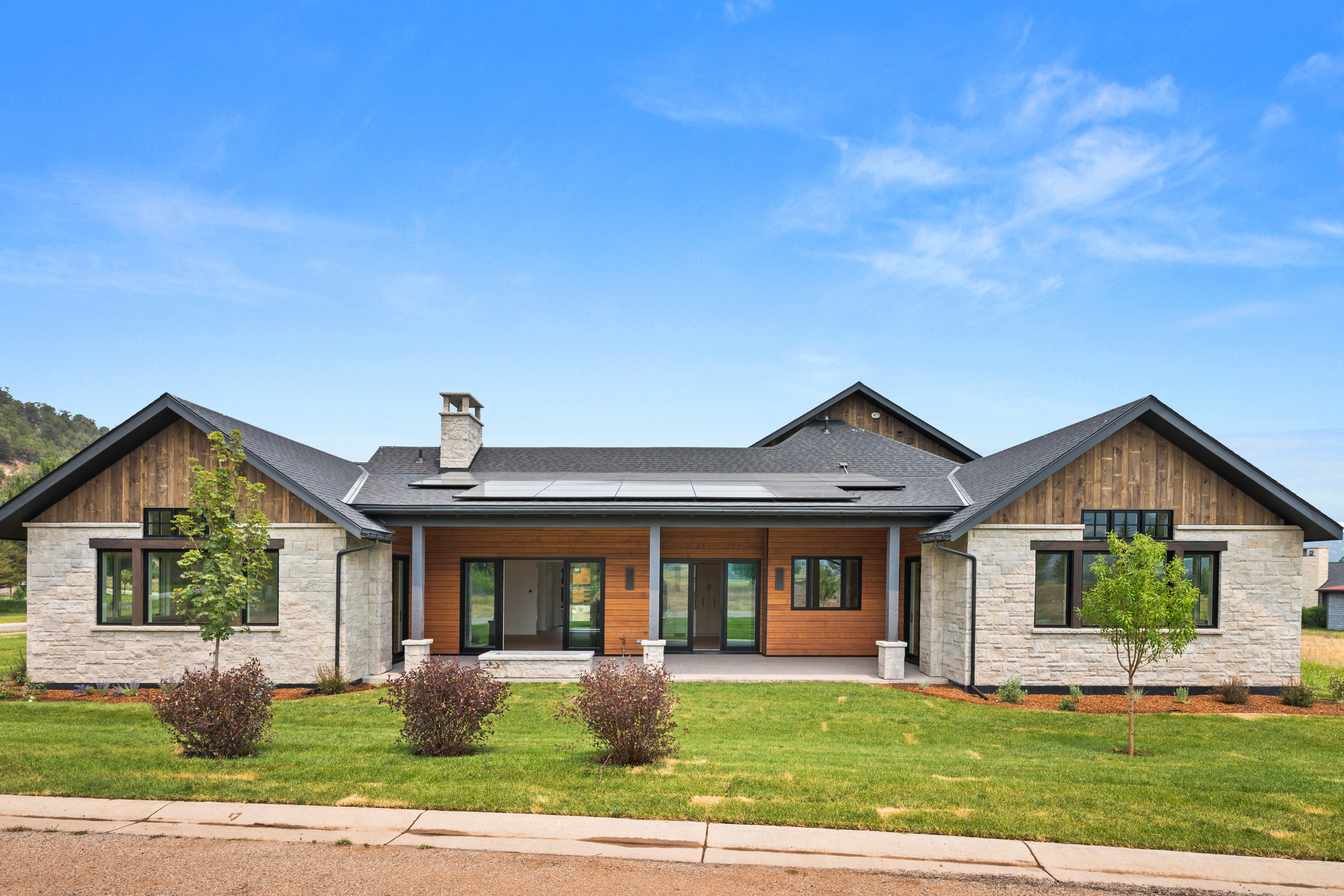 This modern farmhouse features a beautiful mix of textured natural stone and horizontal wood siding, creating an inviting and contemporary curb appeal. The home boasts a sprawling, symmetrical layout with a covered central patio, large energy-efficient windows, and solar panels integrated into the dark-toned roof. A well-manicured lawn with young trees and neatly placed shrubs completes the polished, upscale exterior.