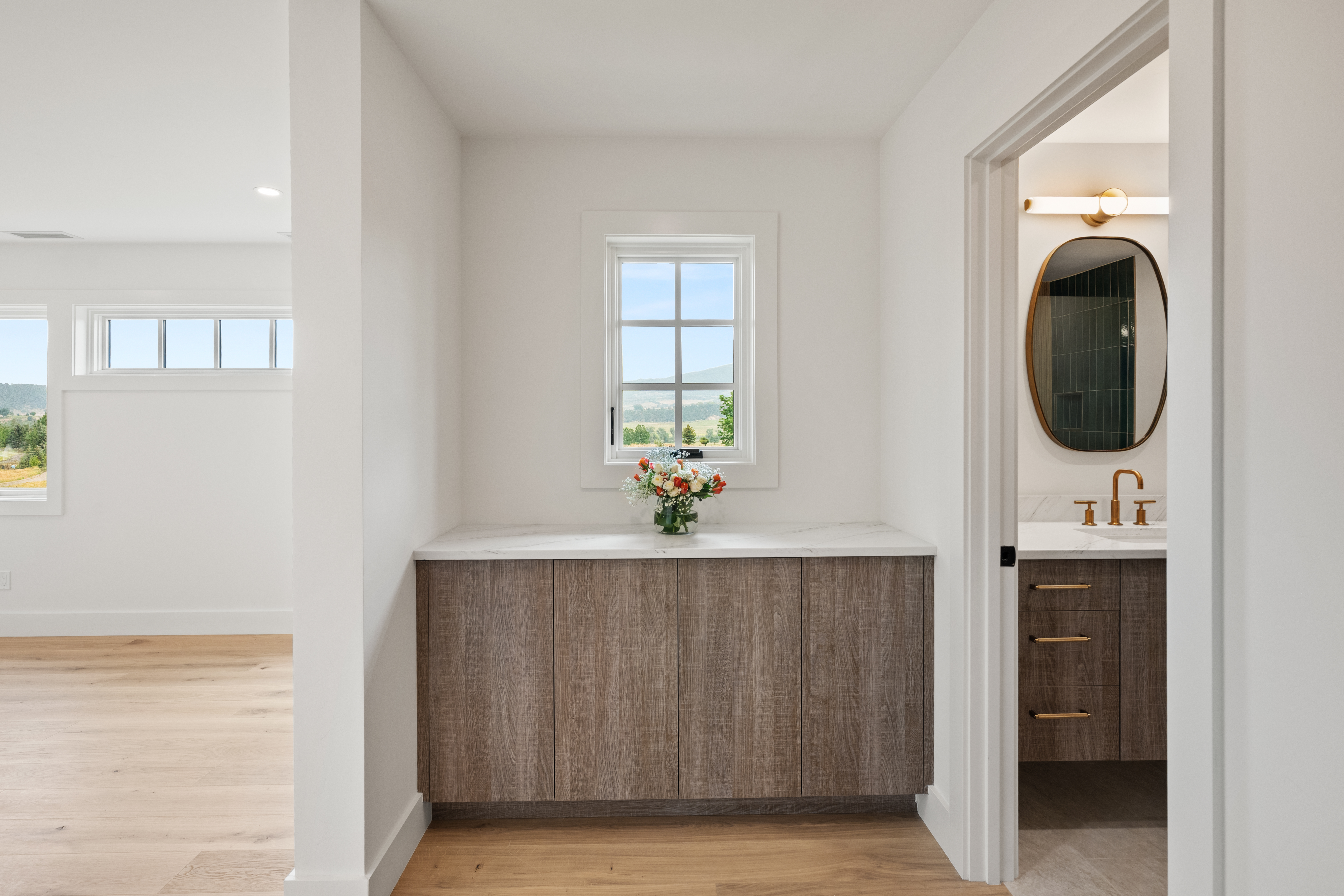 This interior image shows a bright, modern hallway connecting to a bathroom with matching wood-toned cabinetry. The focal point is a built-in console table with a marble top and wood finish, positioned beneath a window that provides natural light and scenic views. The clean lines, light-toned hardwood flooring, and contemporary design elements create a cohesive and welcoming transition space within the home.